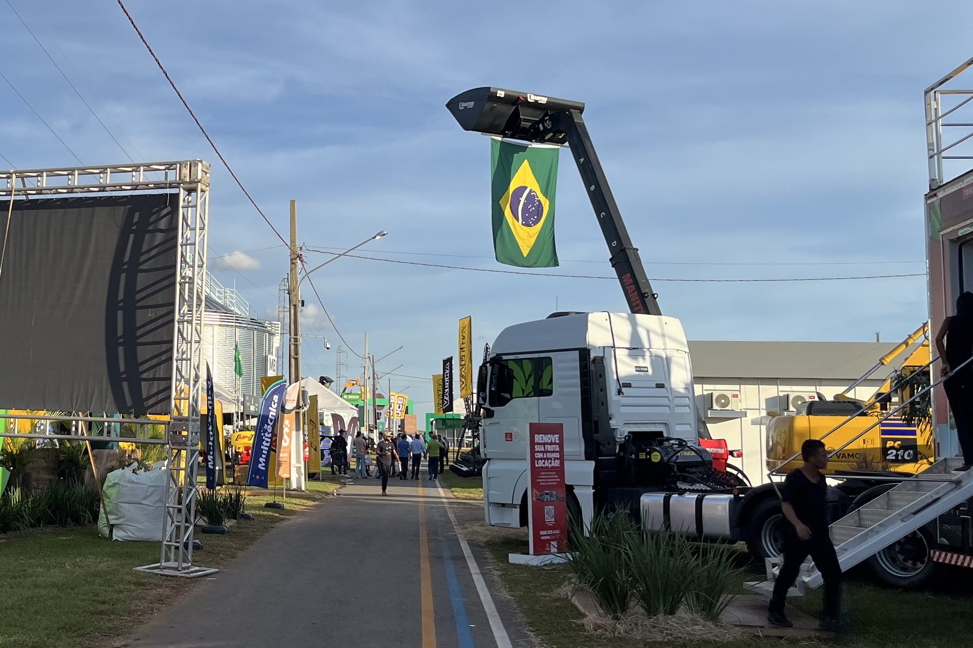 Rua interna do Show Safra BR-163, em Lucas do Rio Verde, com estandes dos dois lados, um silo metálico ao fundo, um caminhão e visitantes caminhando pelo asfalto.