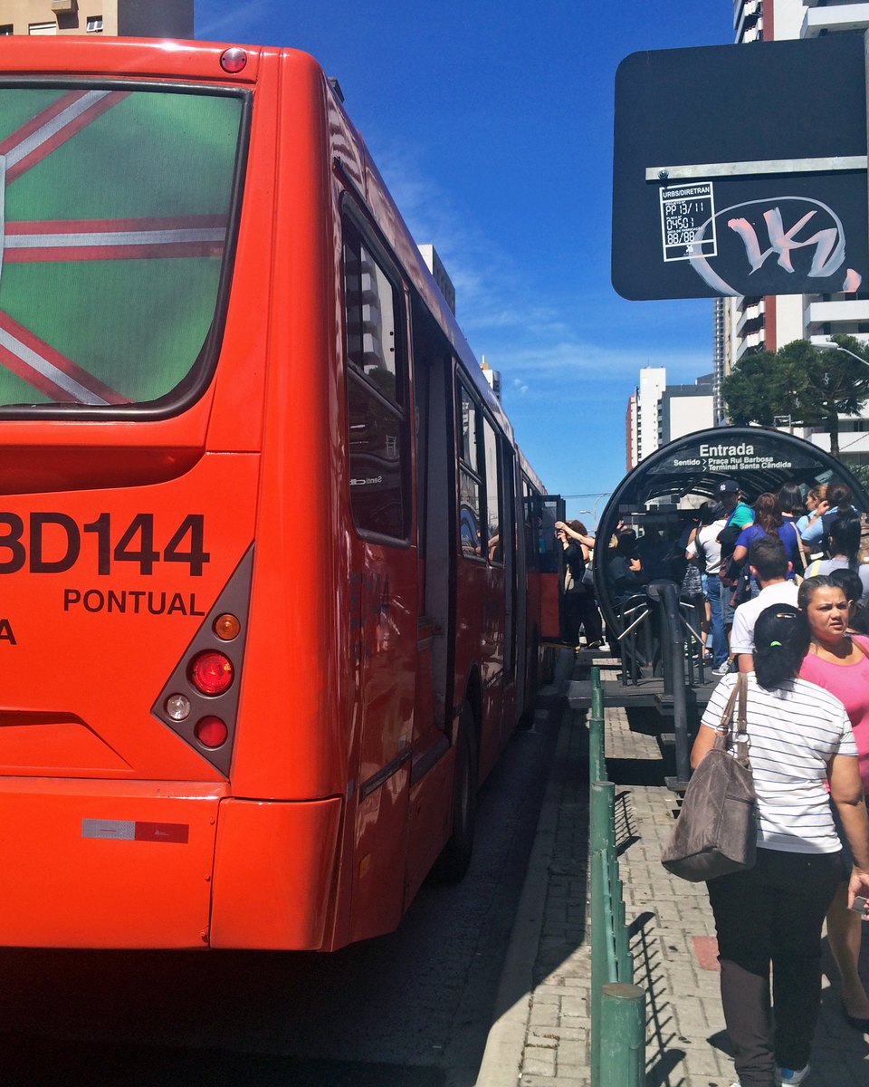 Fila de passageiros embarcando num ônibus vermelho do sistema de Curitiba numa estação-tubo da Avenida Sete de Setembro, num dia de céu azul, com prédios ao fundo.