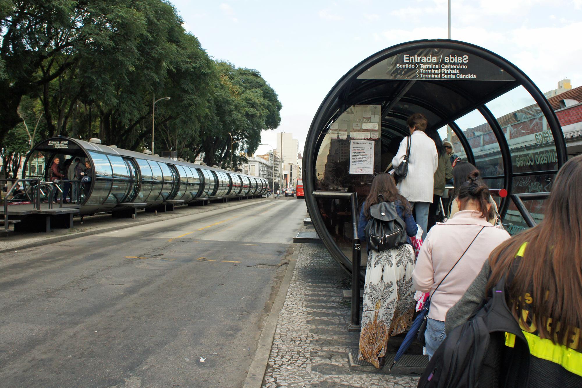 Passageiras com mochila e bolsa entrando numa estação-tubo de ônibus na Praça Eufrásio Correia, em Curitiba, com outra estação-tubo de vidro sob as árvores do outro lado da canaleta exclusiva.