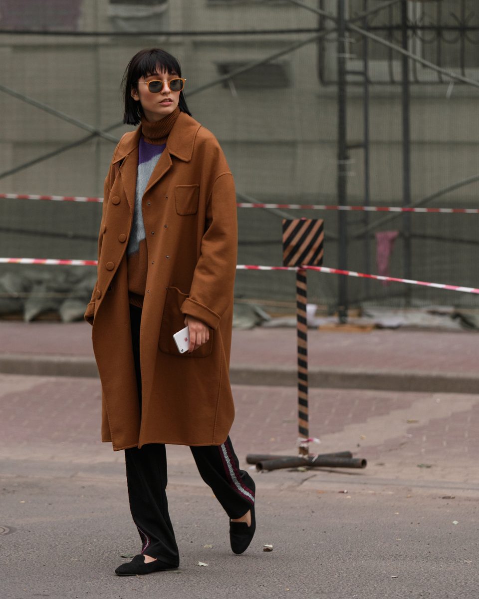 Mulher de casaco caramelo, calça preta e óculos escuros caminhando numa rua, em foto de street style.