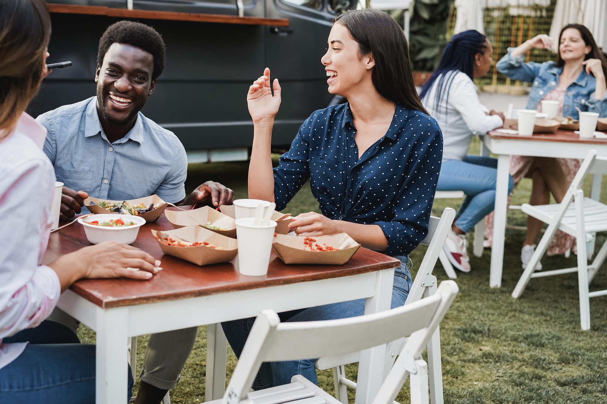 Amigos rindo e comendo em bandejas de papel numa mesa de madeira ao ar livre, sobre a grama, com um food truck escuro ao fundo e outras mesas ocupadas.