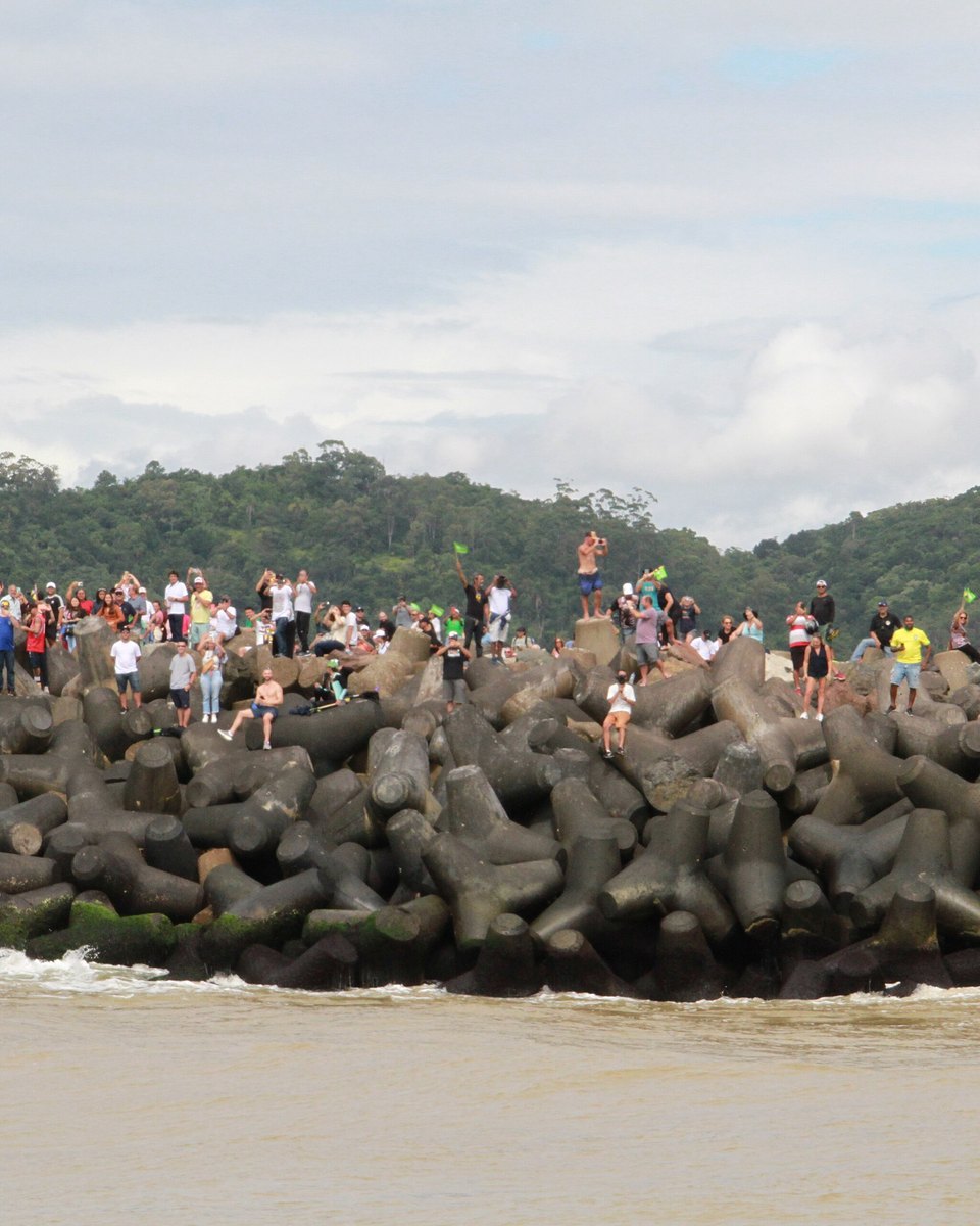 Público em pé sobre os blocos de concreto do molhe da barra do rio Itajaí-Açu, filmando com o celular e acenando para a largada dos veleiros da The Ocean Race em 2023, com o farol listrado de verde e branco à direita e o morro coberto de mata ao fundo.