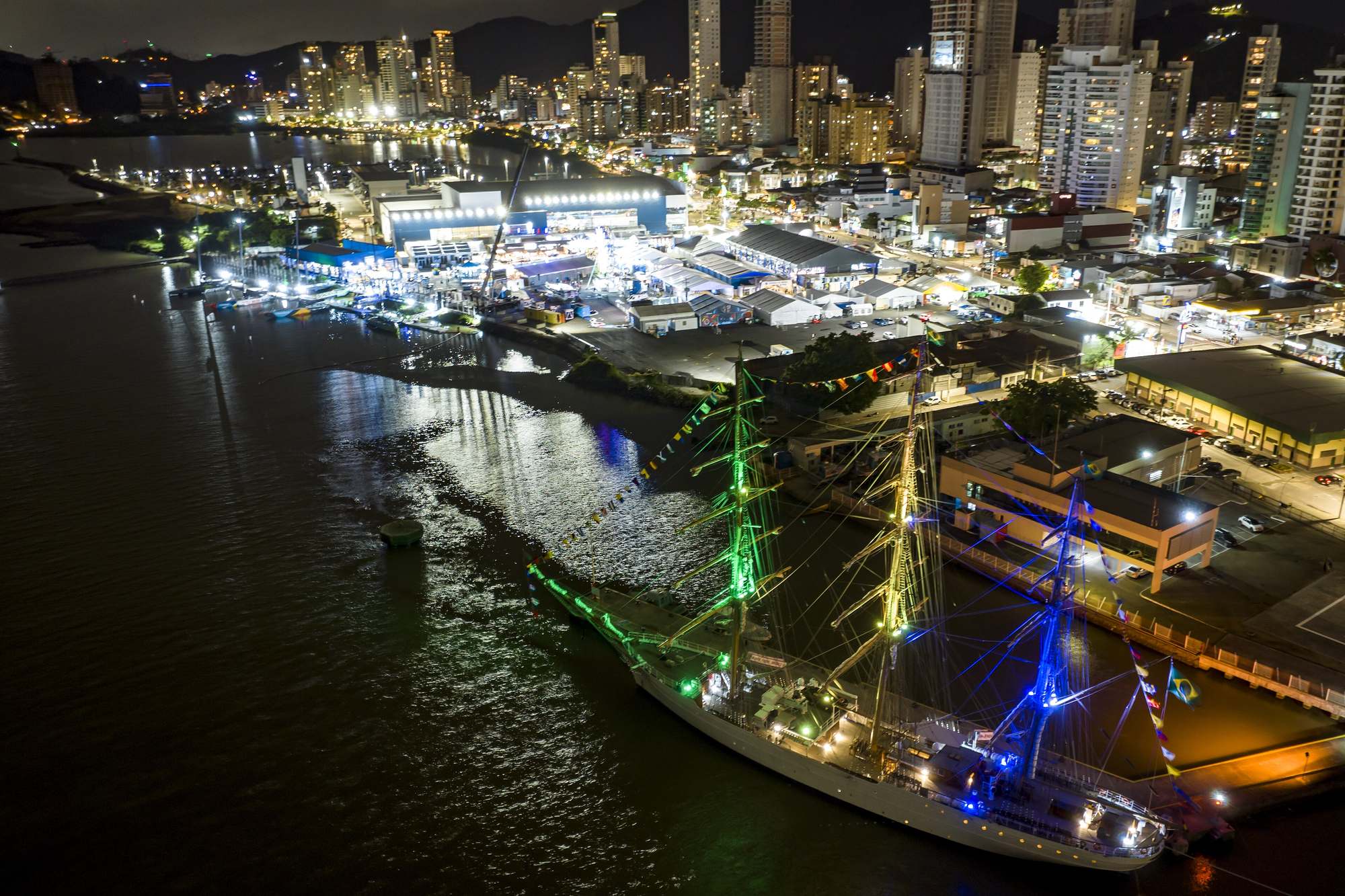 Vista aérea noturna da vila da regata da The Ocean Race em Itajaí, em 2023, com as tendas iluminadas ao lado da marina, o navio-veleiro Cisne Branco atracado com os mastros acesos em verde, amarelo e azul e os prédios da cidade ao fundo.