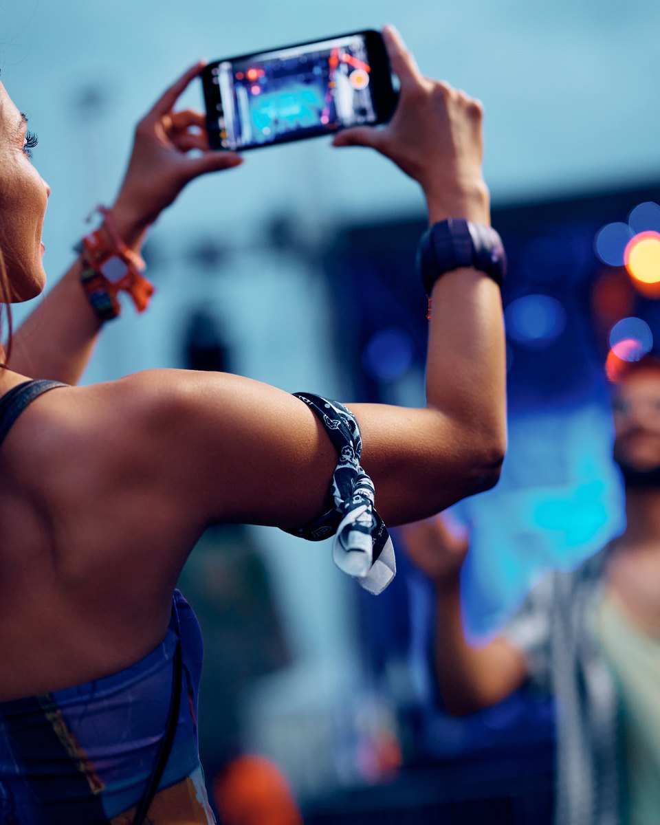 Mulher de faixa trançada no cabelo filmando um amigo com o celular num festival de música, com as luzes do palco desfocadas ao fundo.
