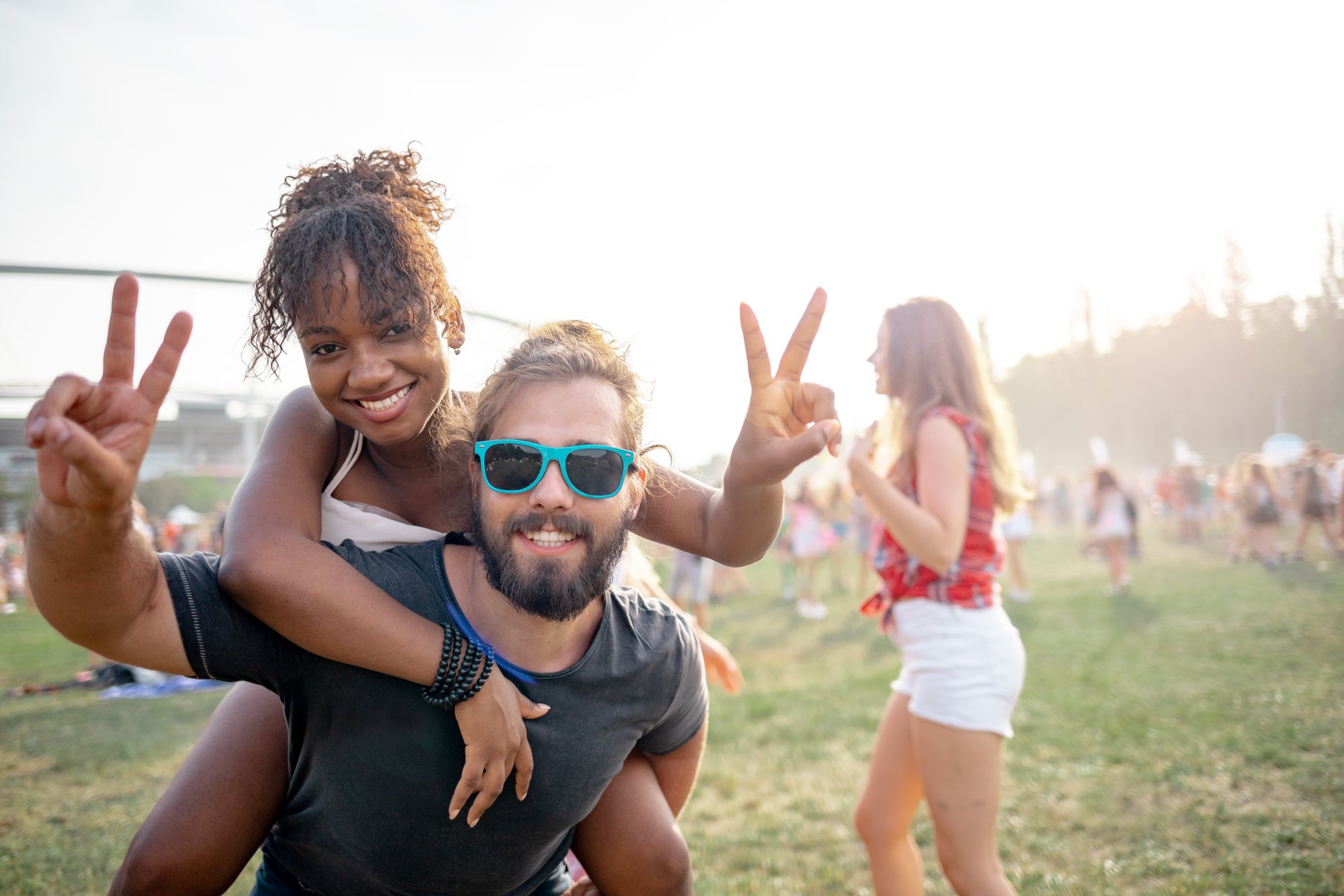 Casal sorrindo e fazendo sinal de paz num festival de música ao ar livre, de dia, ela nas costas dele, com gente andando na grama ao fundo.