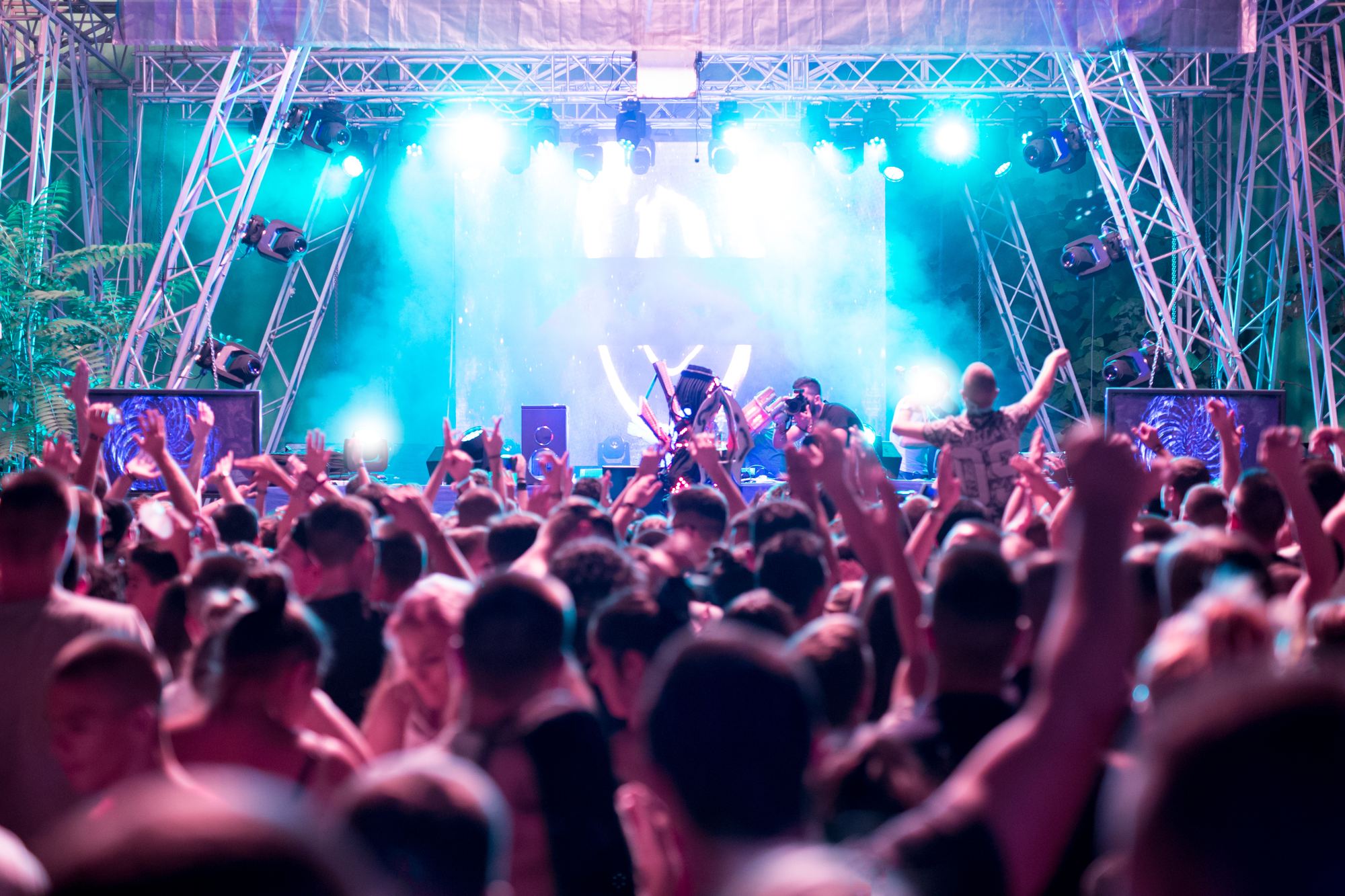 Público com os braços para cima diante de um palco de música eletrônica ao ar livre, com luzes azuis, fumaça e um DJ ao fundo.