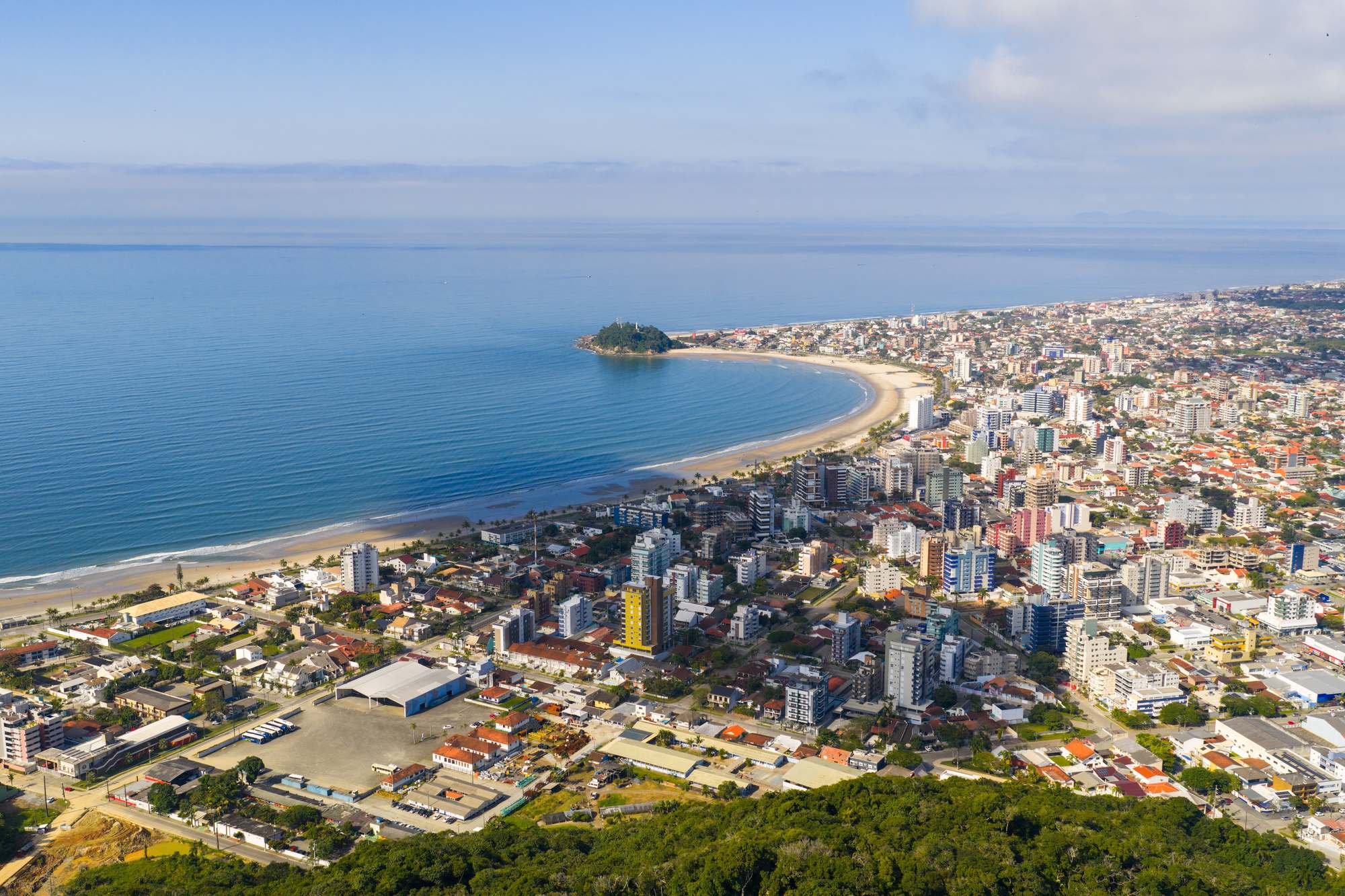 Vista aérea de Guaratuba, no litoral do Paraná, com a faixa de areia em curva, uma ilhota verde na ponta da praia, prédios da orla e o mar calmo ao fundo.