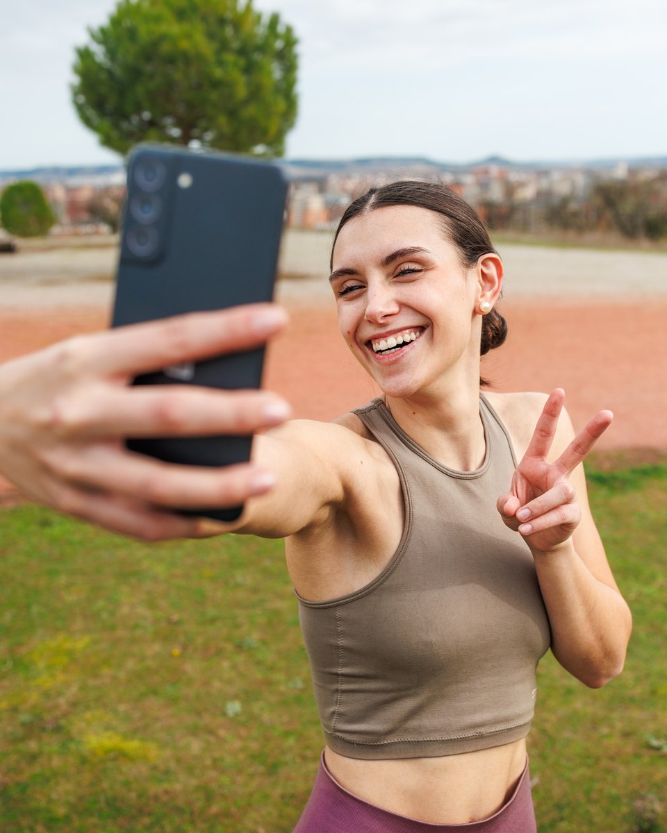 Mulher sorridente de top esportivo e legging tira uma selfie com o celular depois de correr ao ar livre, fazendo sinal de paz com a mão.