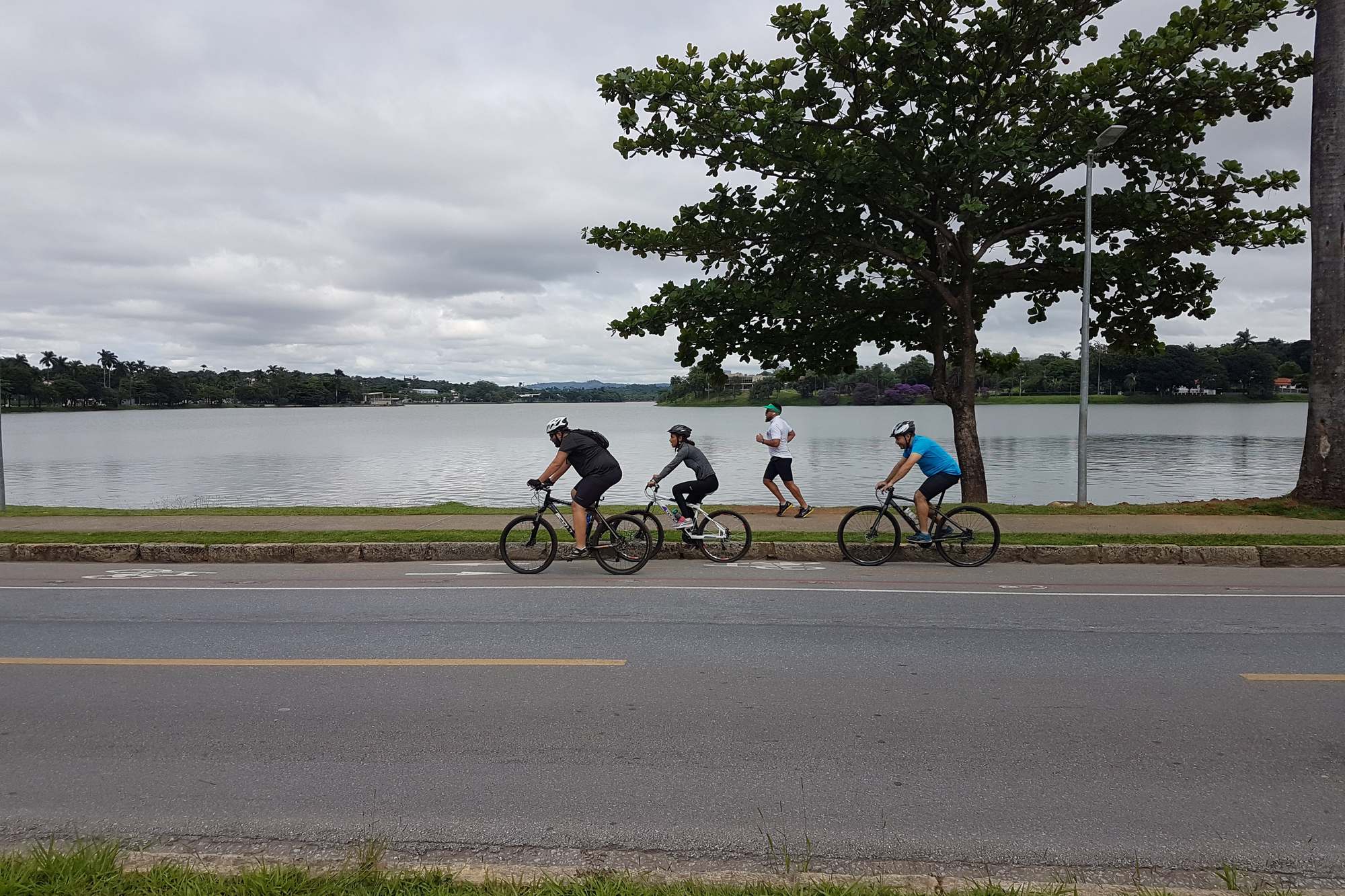 Três ciclistas de capacete e um corredor de camiseta branca na orla da Lagoa da Pampulha, em Belo Horizonte, com a água e uma árvore grande ao fundo, numa manhã nublada.