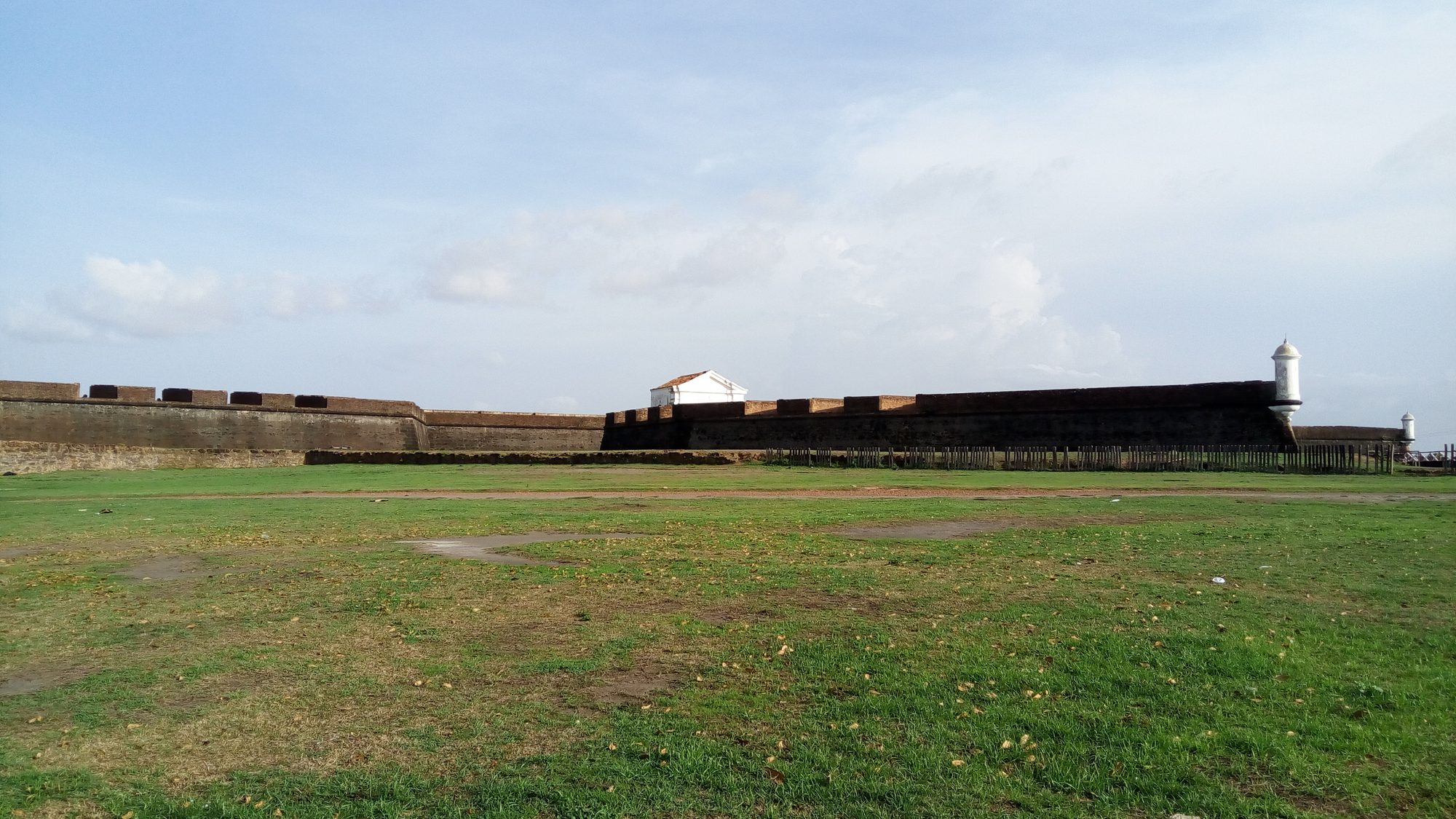 Muralha de pedra e alvenaria da Fortaleza de São José de Macapá vista de fora, com gramado em primeiro plano, uma garita branca ao fundo e céu azul