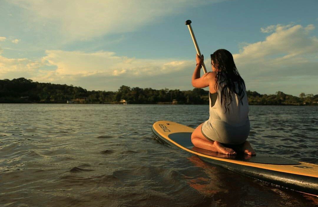 Mulher de costas em pé sobre uma prancha de stand up paddle no rio Araguari ao entardecer, com a mata na margem e o céu em tons dourados