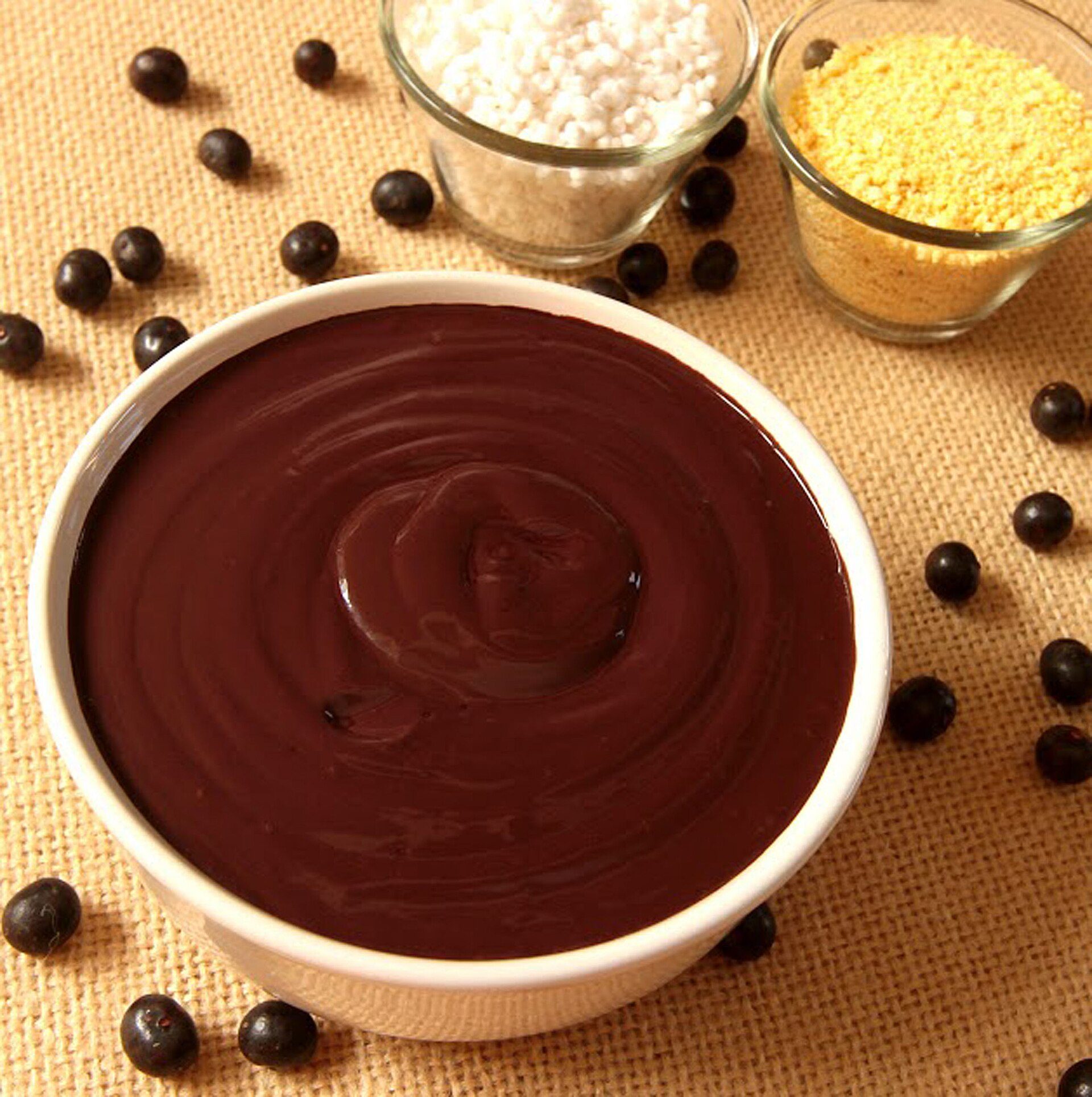 Tigela de açaí batido, roxo e cremoso, cercada por frutas de açaí espalhadas, farinha de tapioca e farinha de mandioca em potes de vidro