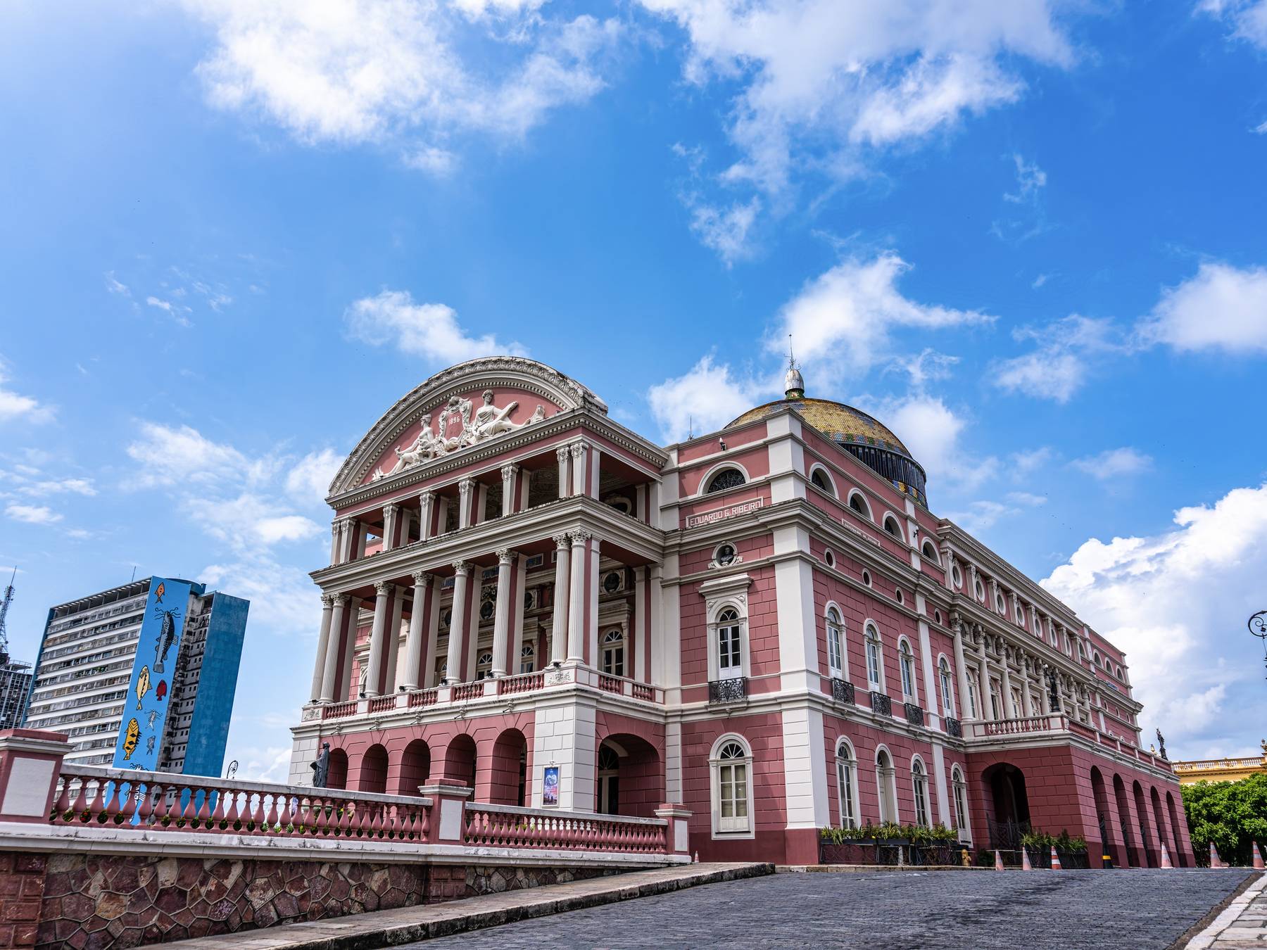 Fachada do Teatro Amazonas em Manaus, com as colunas do pórtico, a cúpula de mosaico colorido e o céu azul com nuvens brancas atrás