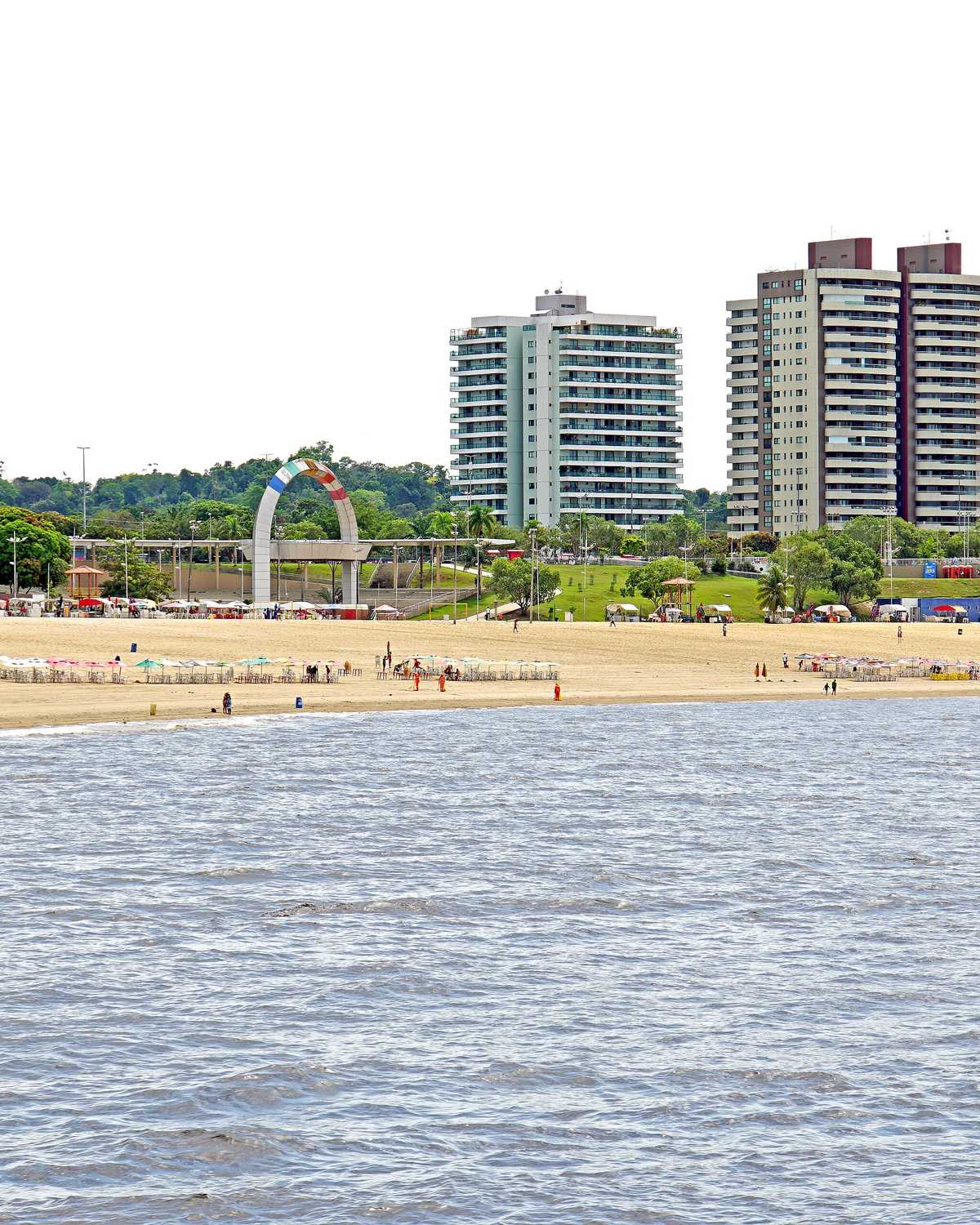 Praia da Ponta Negra, em Manaus, vista do rio Negro, com faixa de areia cheia de guarda-sóis e banhistas e os prédios do bairro ao fundo.