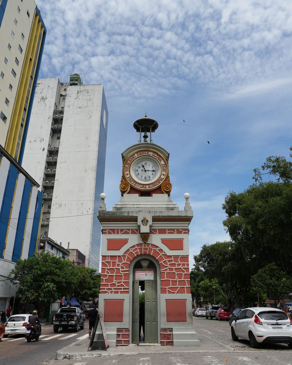 Relógio Municipal no calçadão da Avenida Eduardo Ribeiro, no centro de Manaus, com lojas, carros estacionados e pedestres passando na calçada.