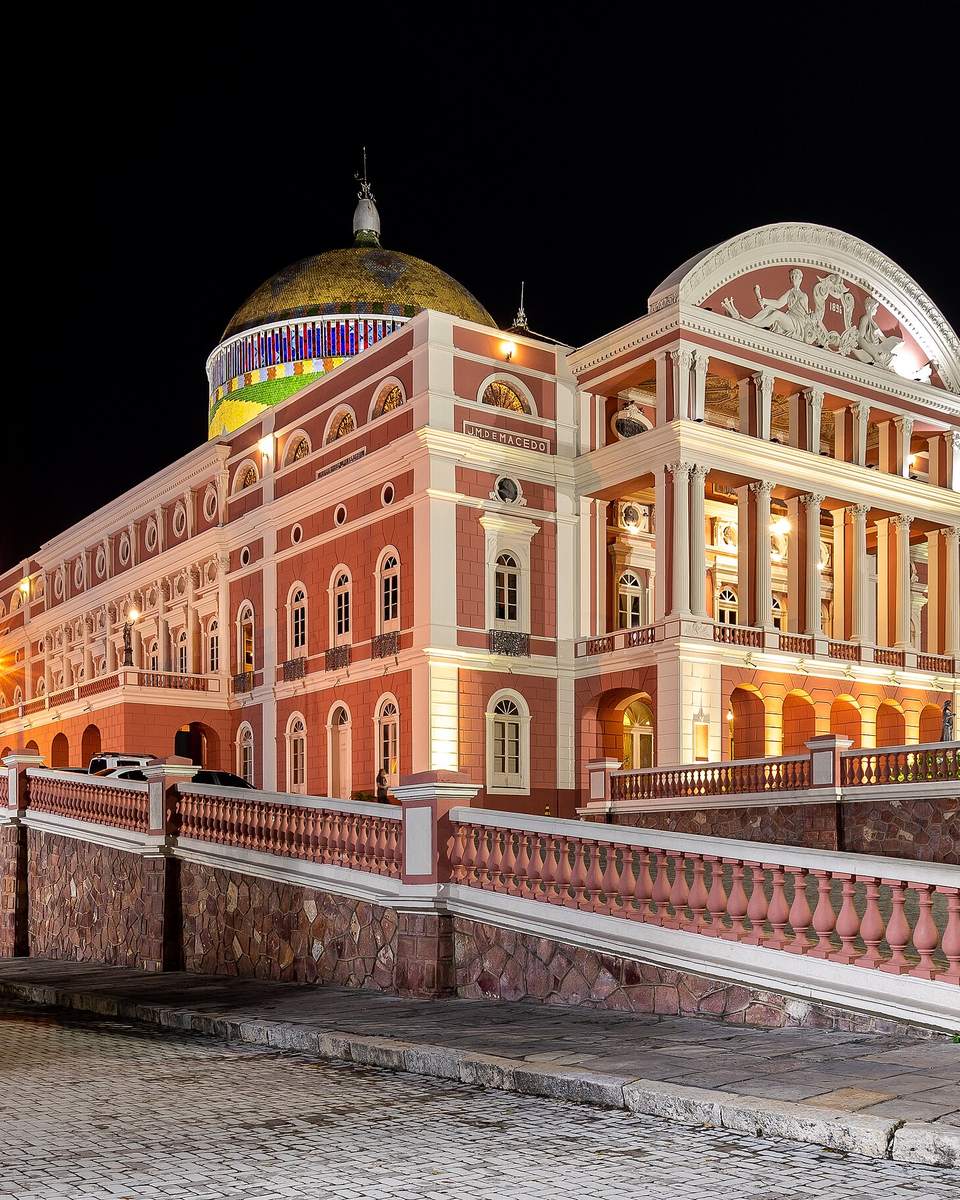 Teatro Amazonas iluminado à noite em Manaus, visto da esquina da praça, com a fachada rosa e branca acesa e a cúpula de mosaico colorido no alto.