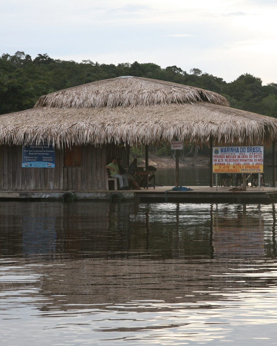 Casa flutuante de madeira com telhado de palha ancorada num braço do rio Negro, perto de Manaus, com hóspedes sentados no deque e a floresta fechada na margem ao fundo.