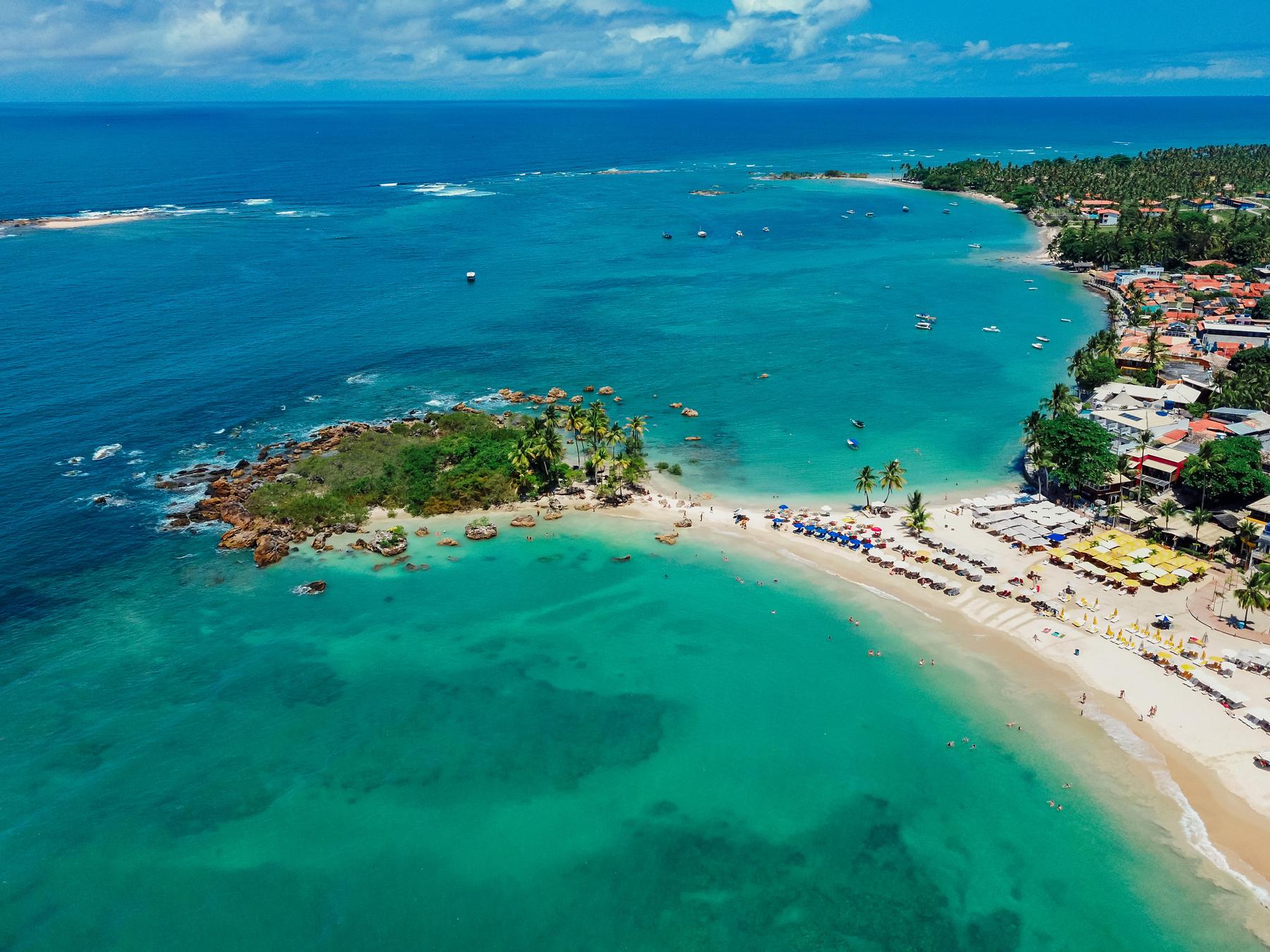 Vista aérea de Morro de São Paulo, com a ilhota coberta de coqueiros ligada à praia, o mar azul-turquesa, a faixa de areia com guarda-sóis e as pousadas da vila na encosta.