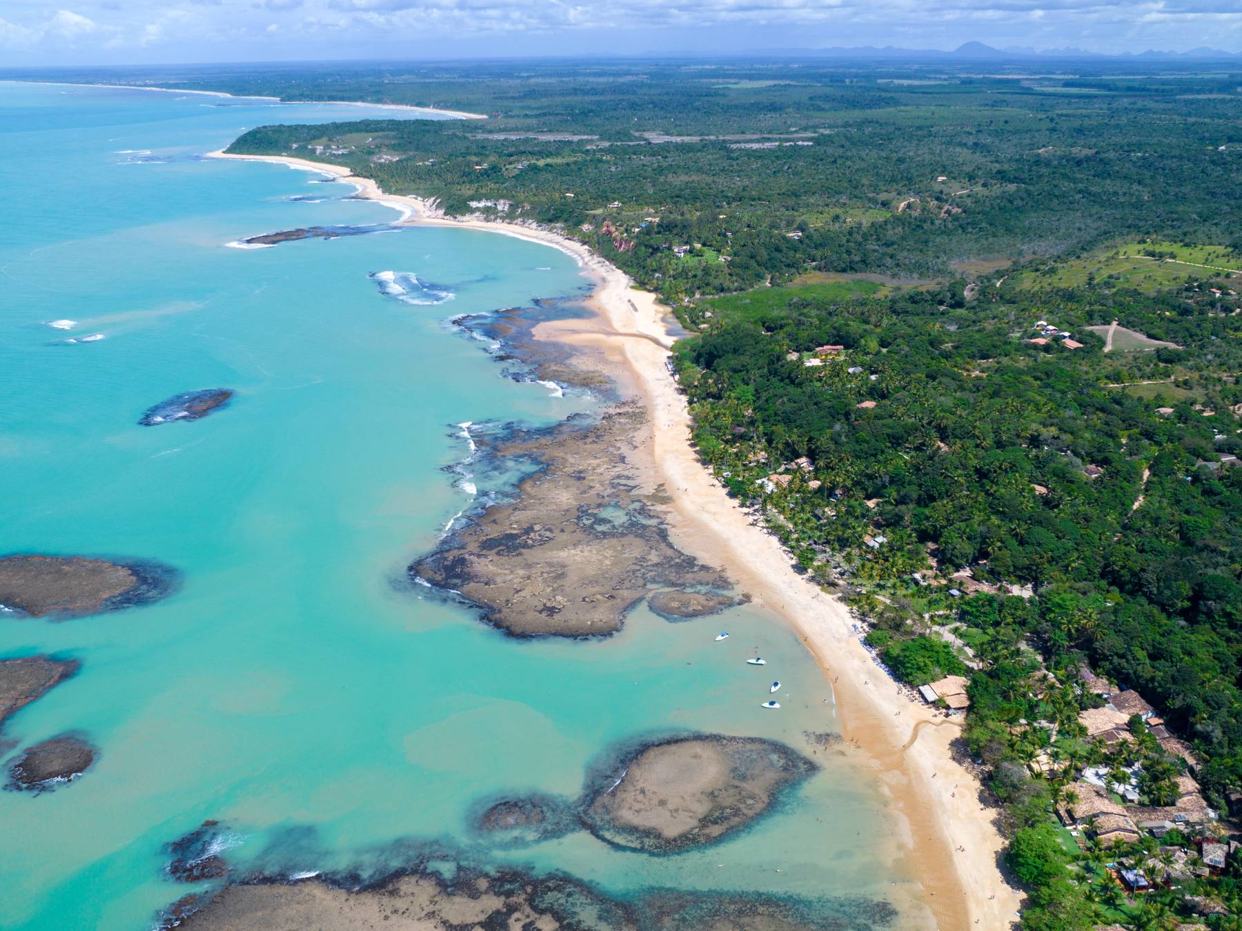 Praia do Espelho vista do alto, com piscinas naturais de recifes no mar verde-claro, a areia dourada e a mata atlântica com casas entre as árvores.