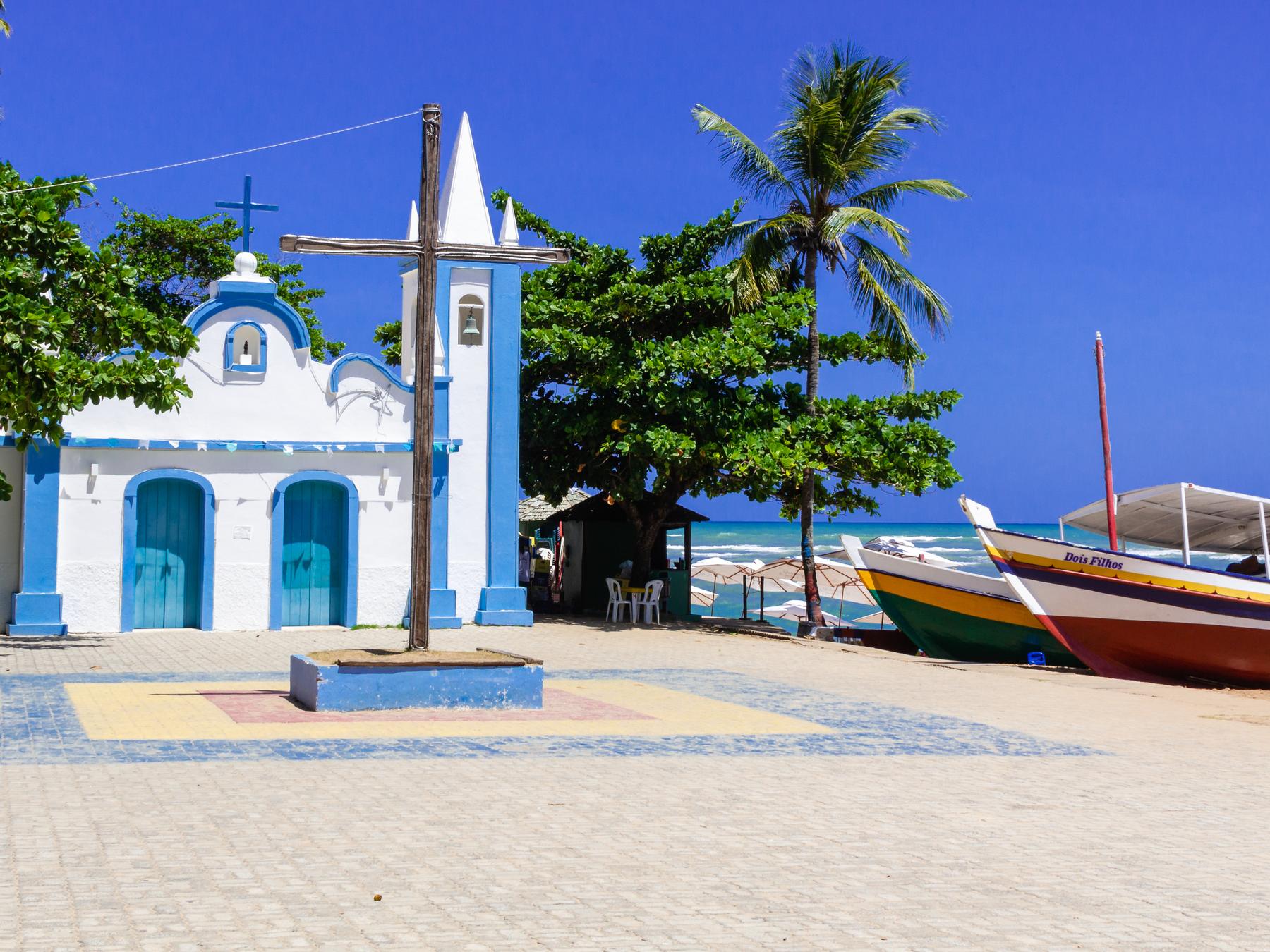 Igrejinha branca e azul de Praia do Forte na praça de pedra, com um cruzeiro, coqueiros, barcos de pesca coloridos na areia e o céu azul.
