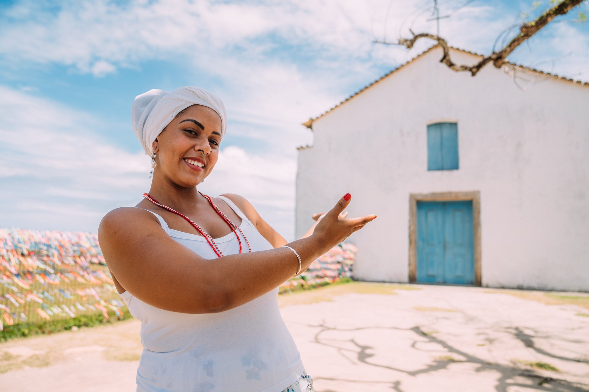 Mulher sorrindo com turbante branco, colar de contas vermelho e roupa branca de baiana, apontando para uma igreja branca de janela azul no Centro Histórico de Porto Seguro, com fitinhas coloridas ao fundo.