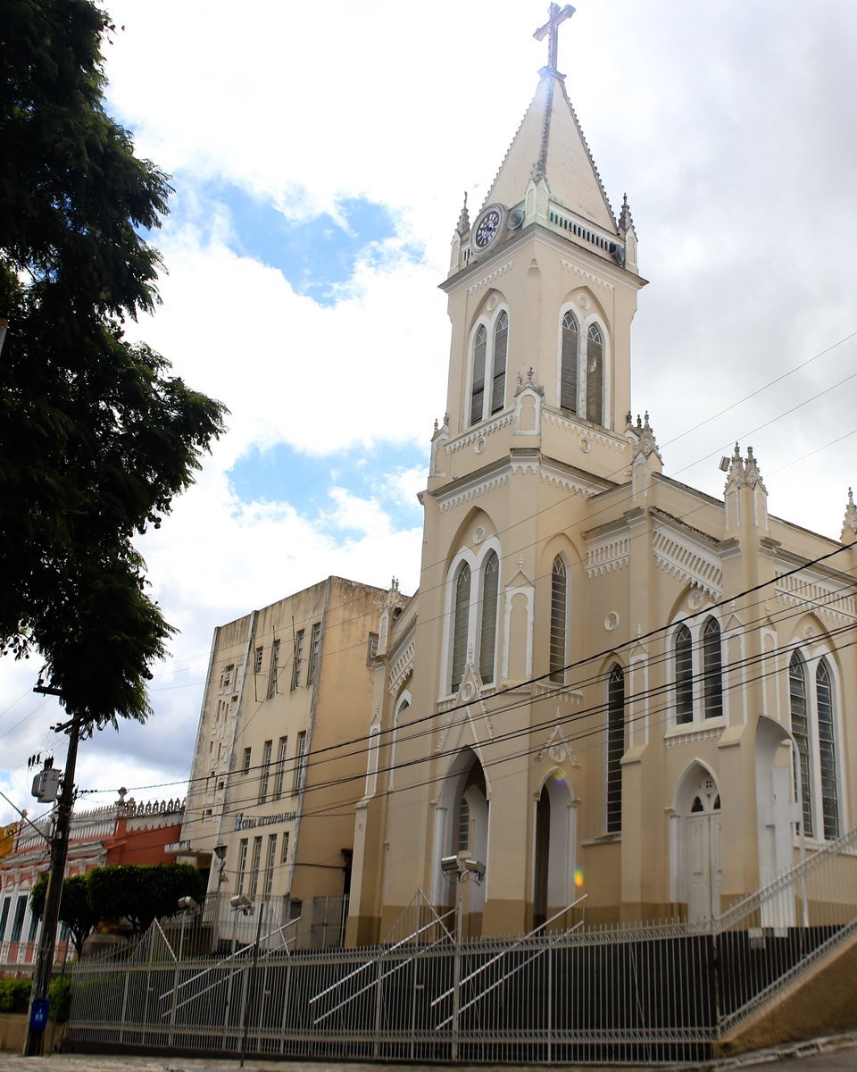 Catedral Nossa Senhora das Vitórias, em Vitória da Conquista, de fachada clara e torre pontiaguda, com a rua em frente e o céu com nuvens.