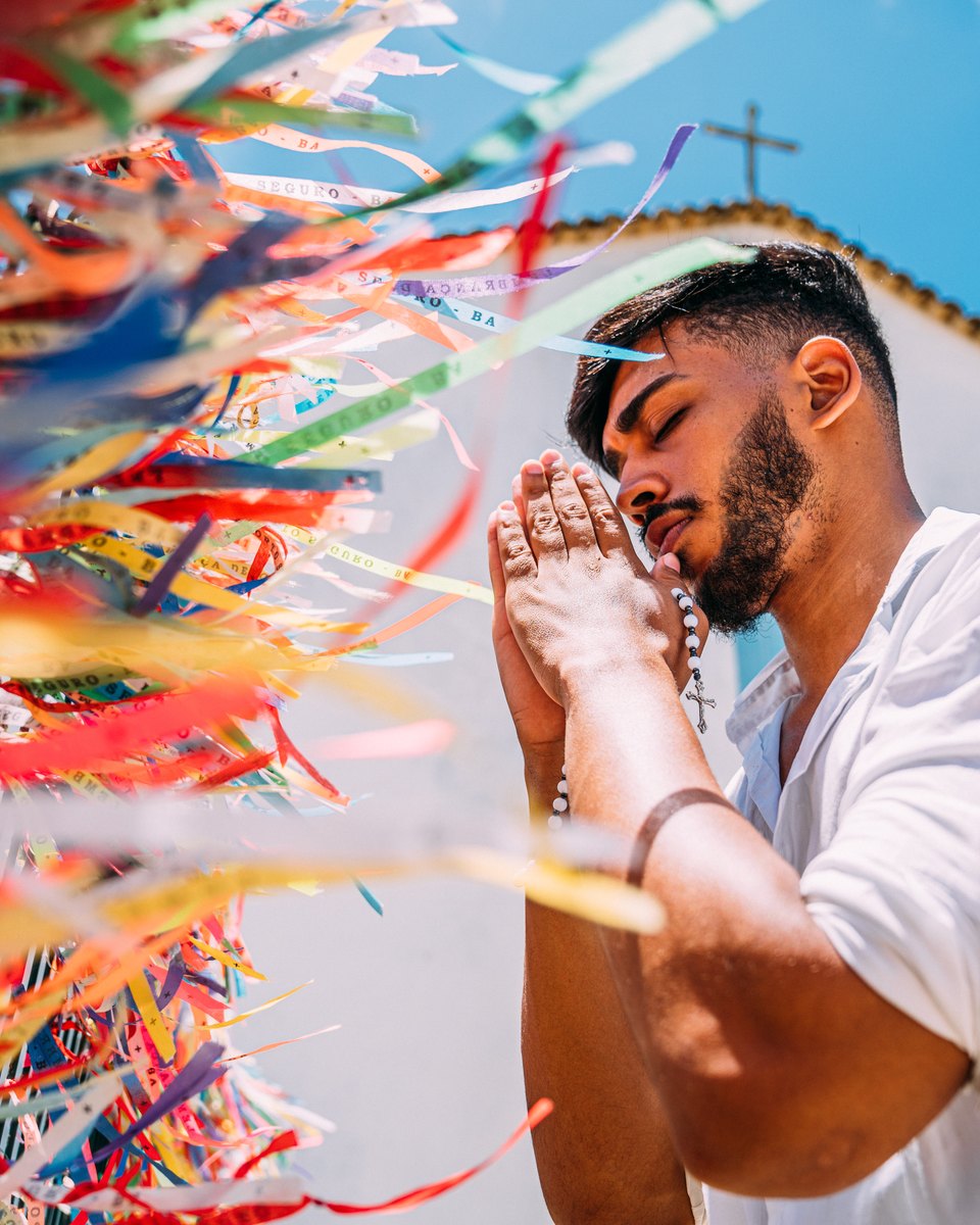 Homem de camisa branca rezando de olhos fechados, com as mãos juntas e um terço, ao lado das fitinhas coloridas amarradas no gradil de uma igreja branca em Arraial d'Ajuda, sob céu azul.