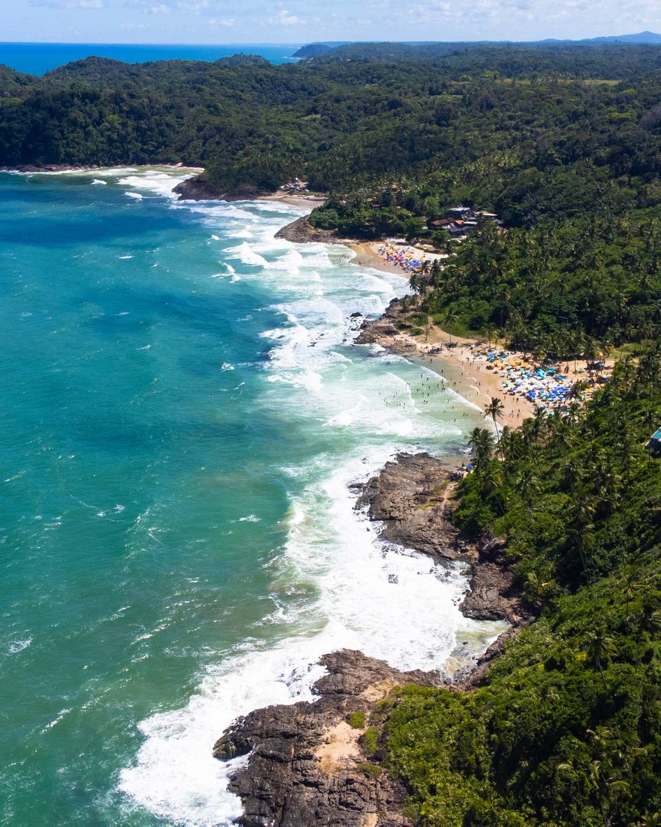 Vista aérea de Itacaré, com o mar verde-azulado, a arrebentação nas pedras, a faixa de areia e a Mata Atlântica com casas entre as árvores.