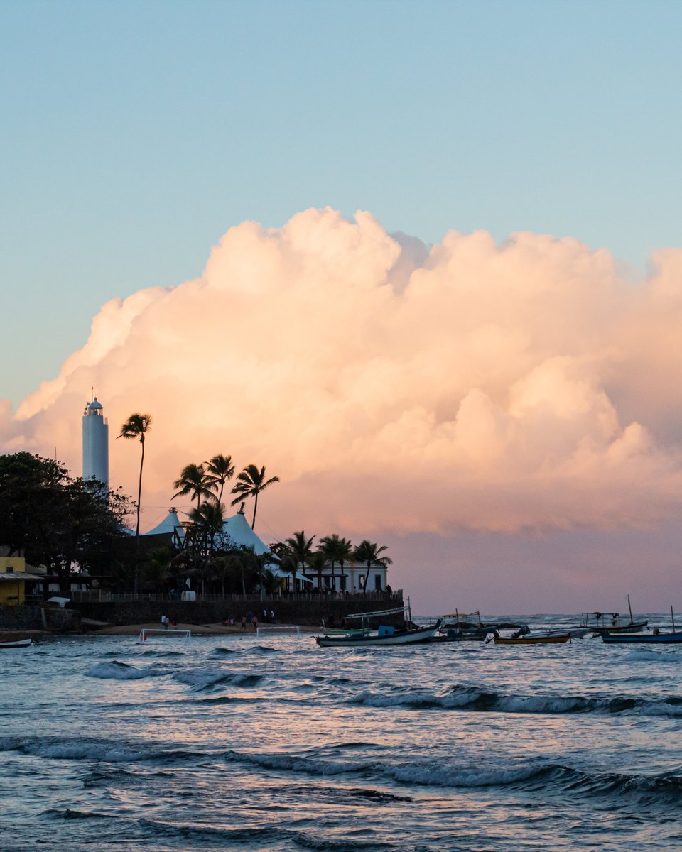Fim de tarde em Praia do Forte, com o farol branco entre coqueiros, barcos de pesca ancorados no mar e uma grande nuvem rosada no céu.