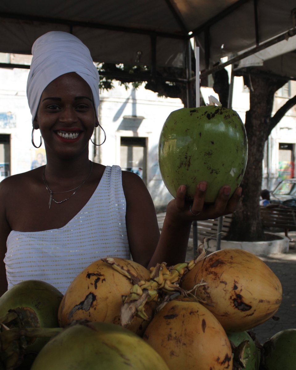 Mulher sorridente de turbante branco segurando um coco verde numa banca de cocos em Cachoeira, com a rua, os carros e o casario ao fundo.