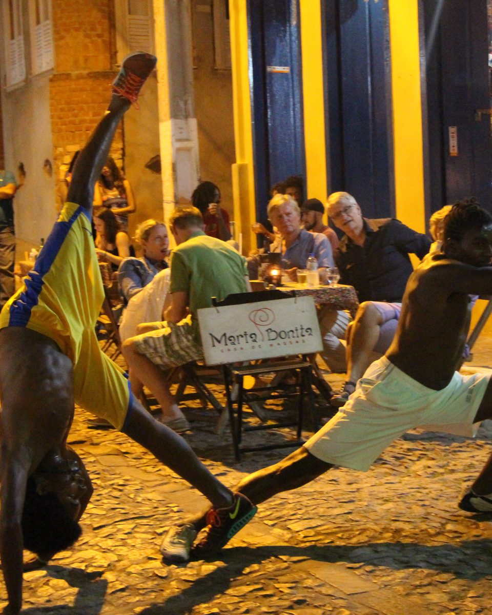 Dois capoeiristas jogando à noite numa rua de pedra de Lençóis, um deles de ponta-cabeça, com moradores e turistas assistindo das mesas em frente aos casarões iluminados.