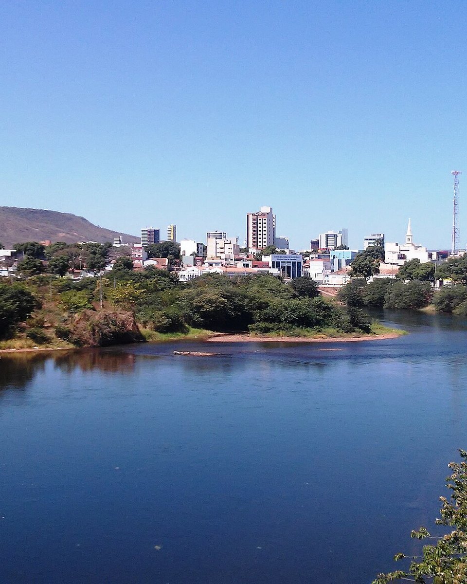Rio Grande, de água azul-escura e margens verdes, atravessando Barreiras, com os prédios da cidade e a serra ao fundo sob céu azul.
