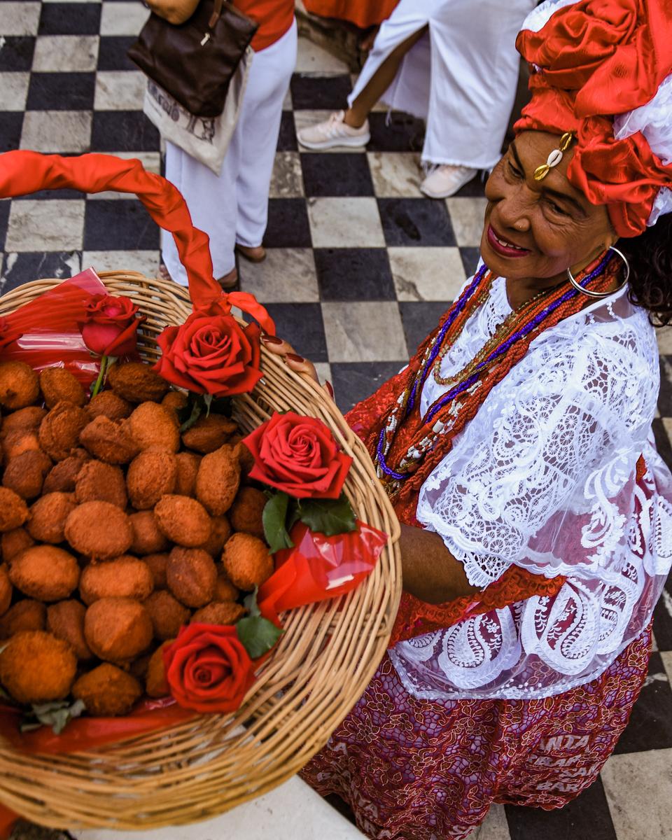 Baiana de turbante vermelho e roupa branca rendada carregando uma cesta de acarajés enfeitada com rosas e fitas vermelhas, sobre um piso de ladrilhos pretos e brancos, durante a Festa de Santa Bárbara.