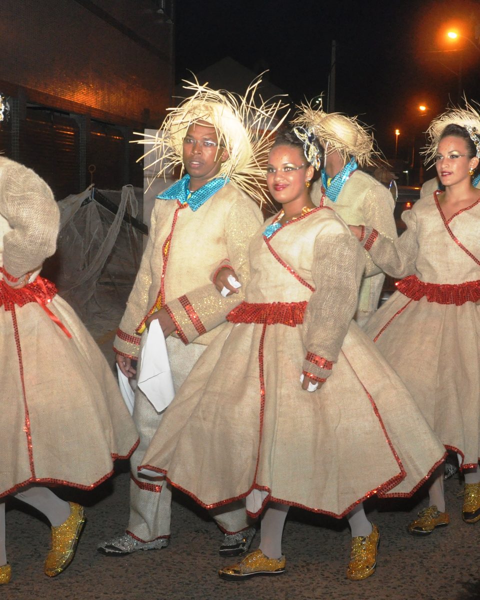 Dançarinas de quadrilha junina com vestidos cor de palha, faixas vermelhas e chapéus de palha desfiada dançando na rua à noite, durante um campeonato de quadrilhas na Bahia.