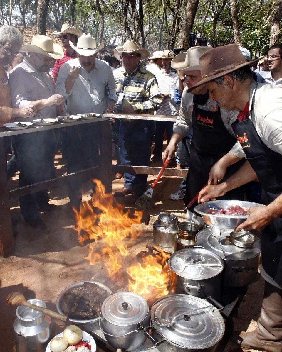 Cozinheiros de comitiva de avental mexendo panelas de alumínio sobre fogo de lenha com chamas altas, no chão de terra, e o público de chapéu olhando por trás de um balcão, na Queima do Alho em Barretos.
