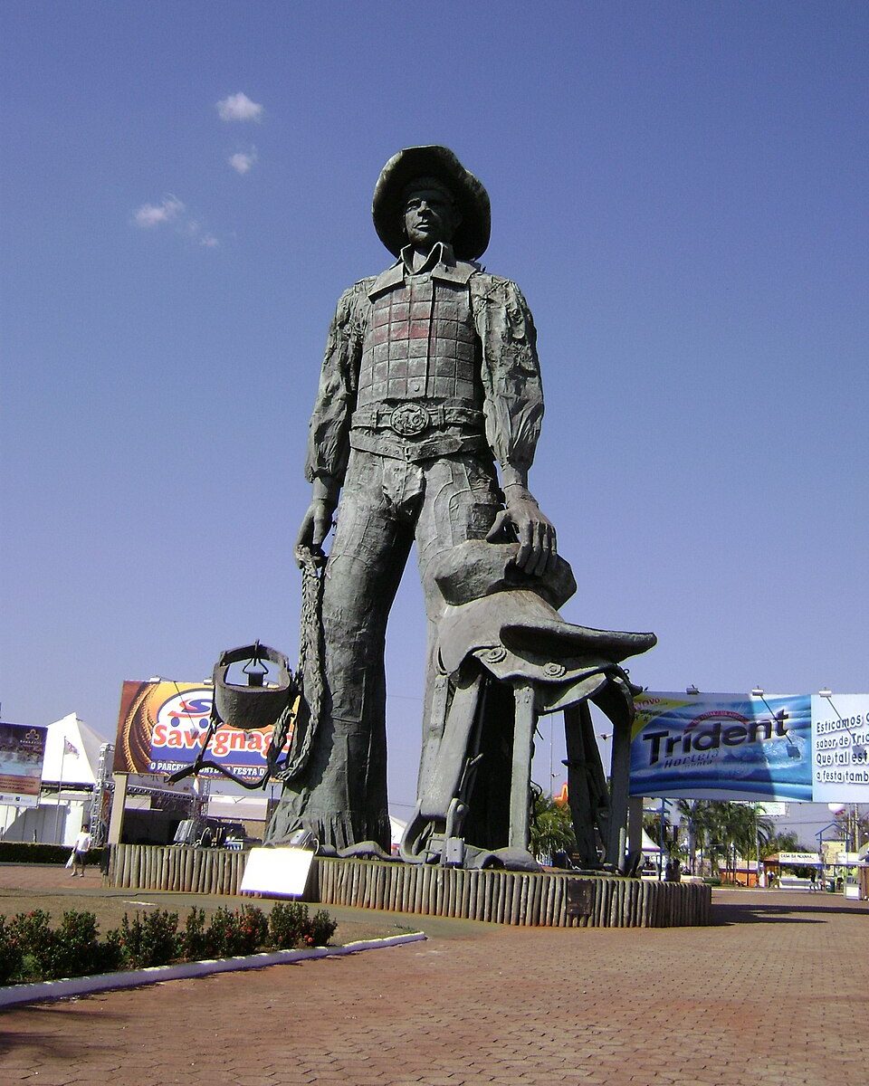 Estátua de um peão de chapéu segurando a sela, sobre um pedestal no Parque do Peão, em Barretos, contra o céu azul.