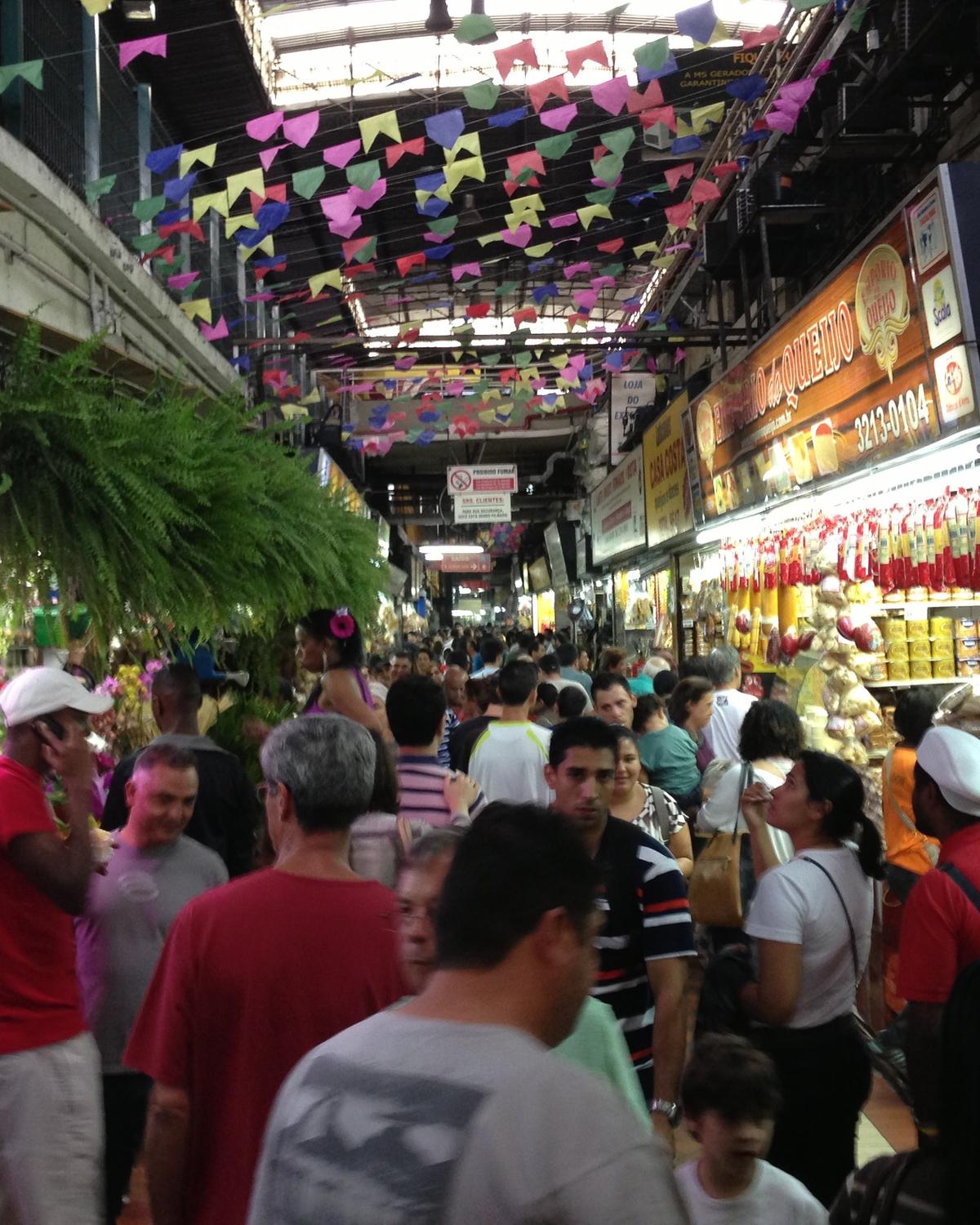 Corredor do Mercado Central de Belo Horizonte lotado de gente entre bancas de flores, artesanato e produtos mineiros, com bandeirinhas coloridas penduradas no teto.