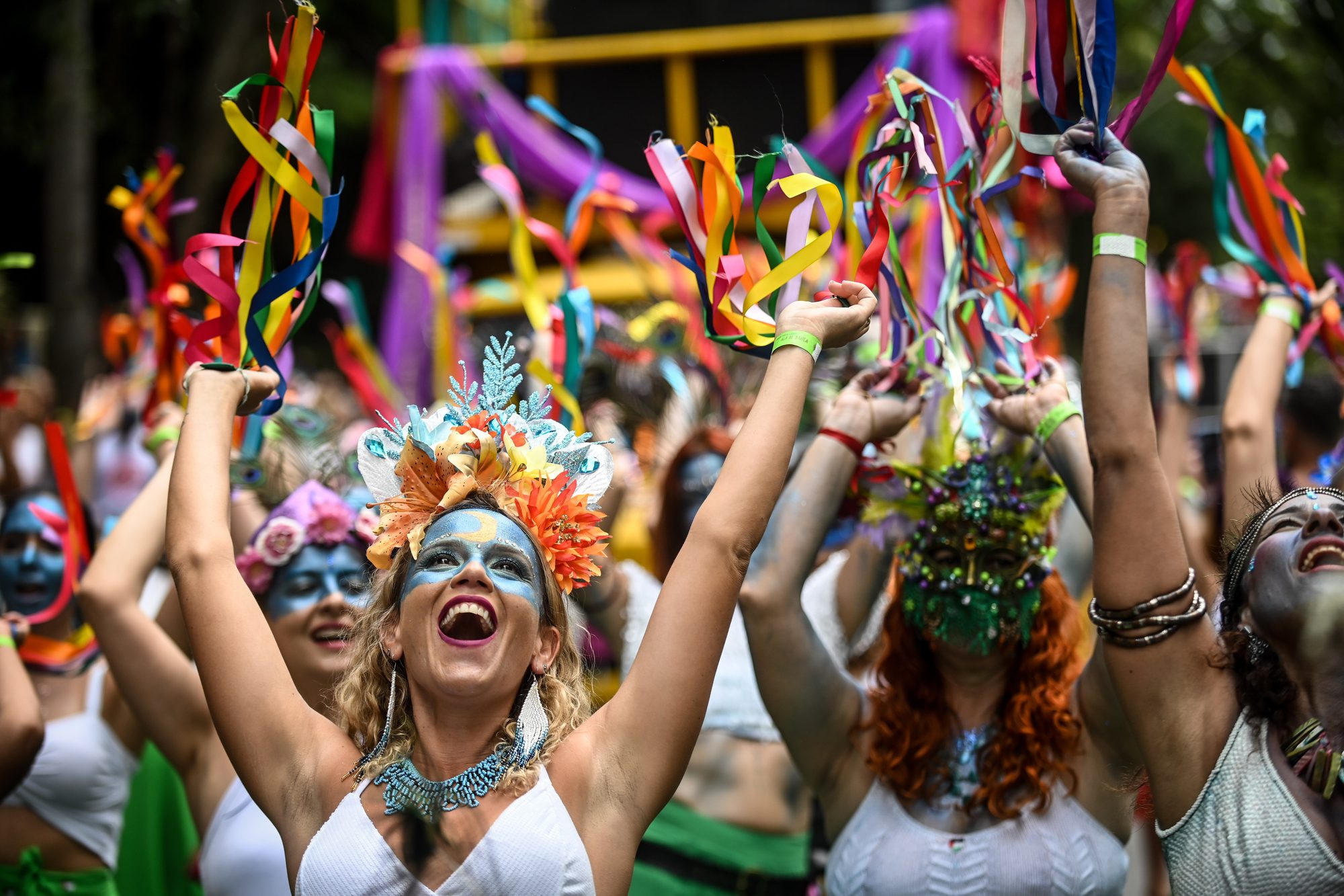 Foliãs fantasiadas com arranjos de flores na cabeça, maquiagem colorida e fitas nas mãos cantando e dançando no desfile do bloco Pena de Pavão de Krishna, no Carnaval de Belo Horizonte.