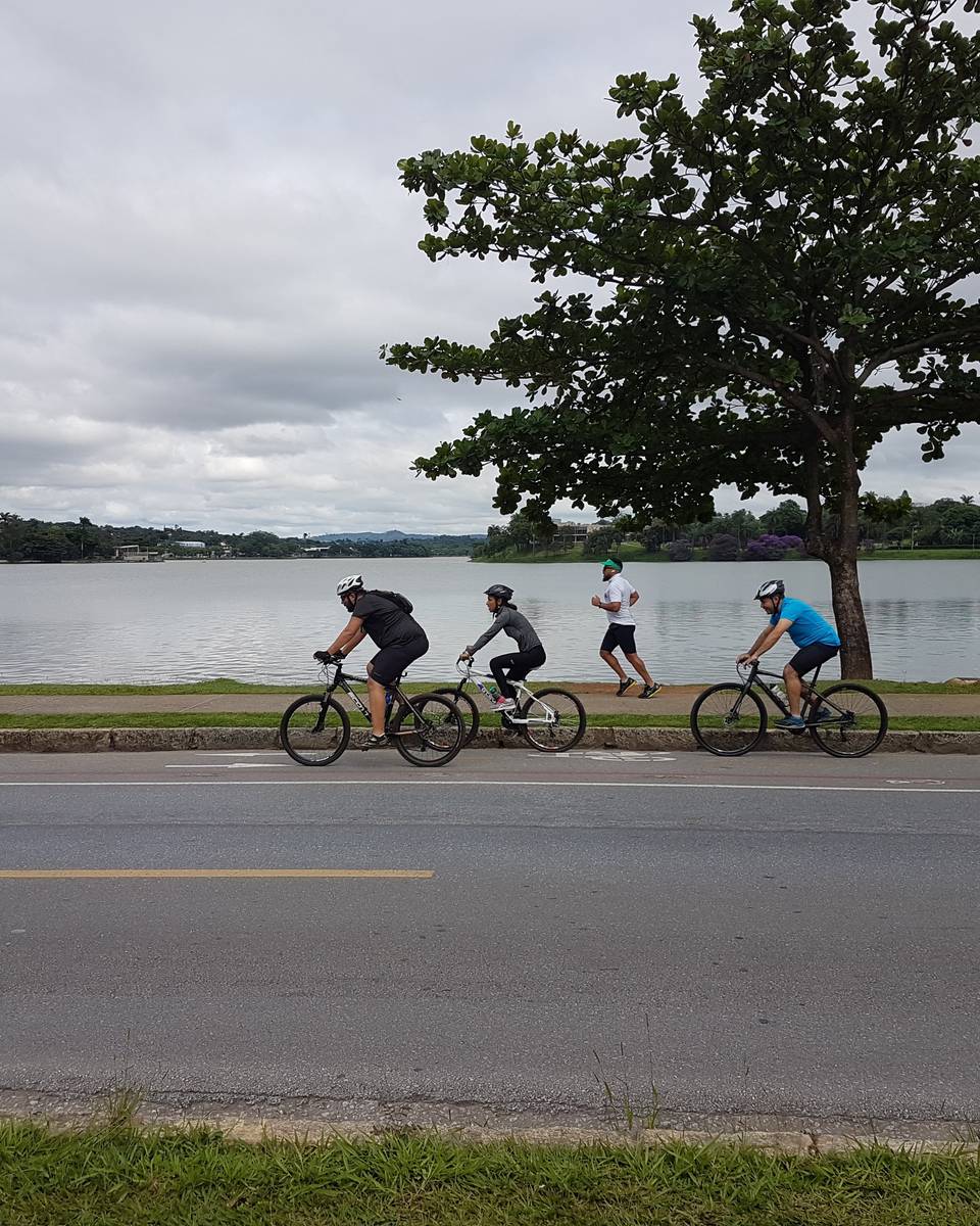 Três ciclistas e um corredor na orla da Lagoa da Pampulha, em Belo Horizonte, num dia nublado, com a água e a vegetação da outra margem ao fundo.