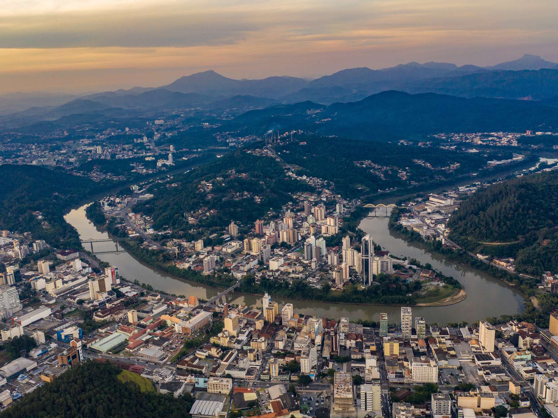 Vista aérea de Blumenau ao entardecer, com o rio Itajaí-Açu fazendo curvas entre os prédios do Centro e os bairros, os morros cobertos de mata e as serras do Vale Europeu no horizonte sob o céu dourado.