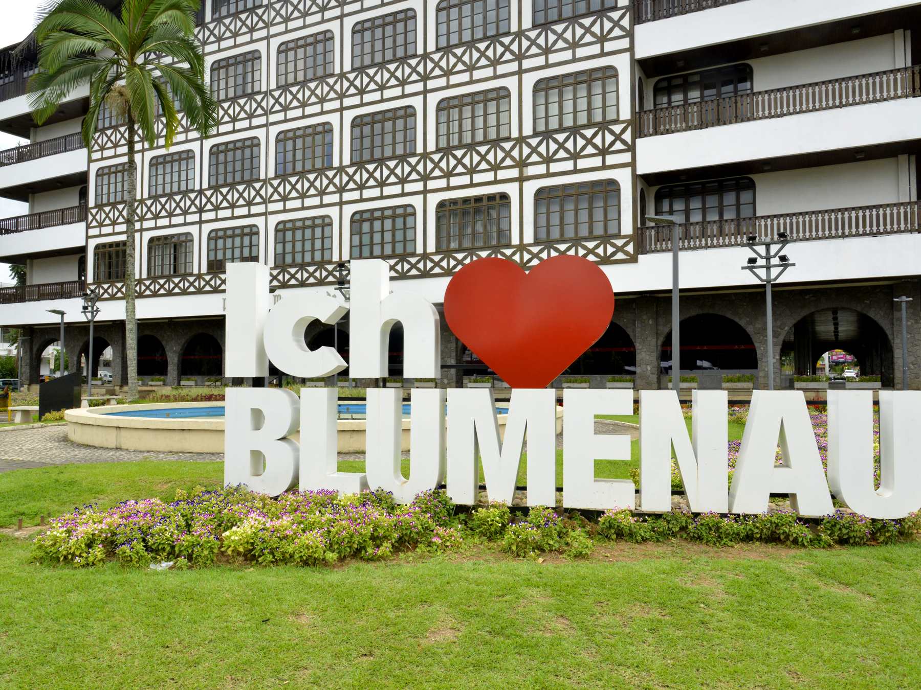 Letreiro branco "Ich liebe Blumenau" com um coração vermelho no gramado florido em frente ao prédio enxaimel da Prefeitura de Blumenau, com palmeiras ao lado.