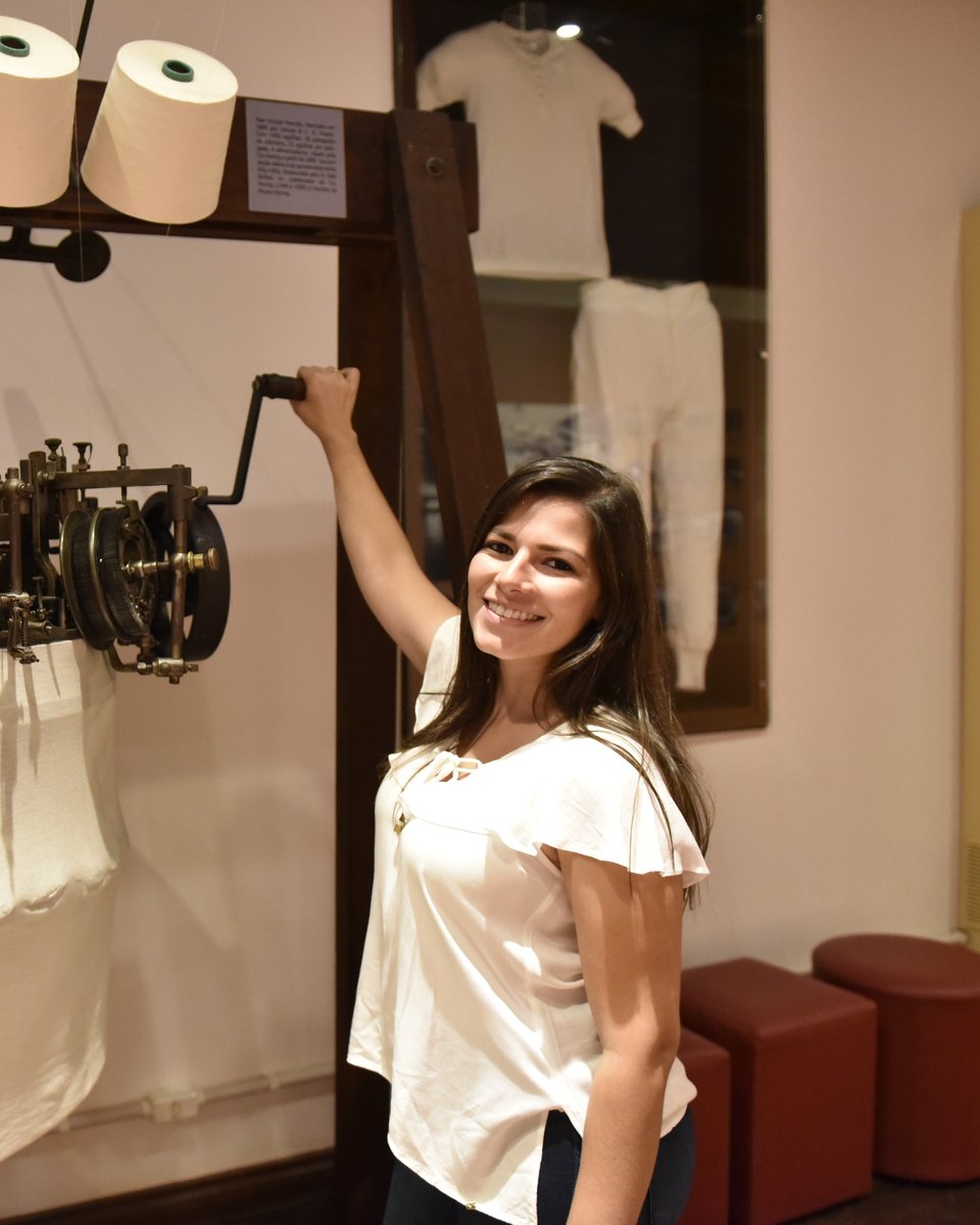 Jovem sorrindo e girando a manivela de uma antiga máquina circular de malharia, com cones de fio no alto, no Museu Hering, em Blumenau, com peças de roupa branca expostas atrás.