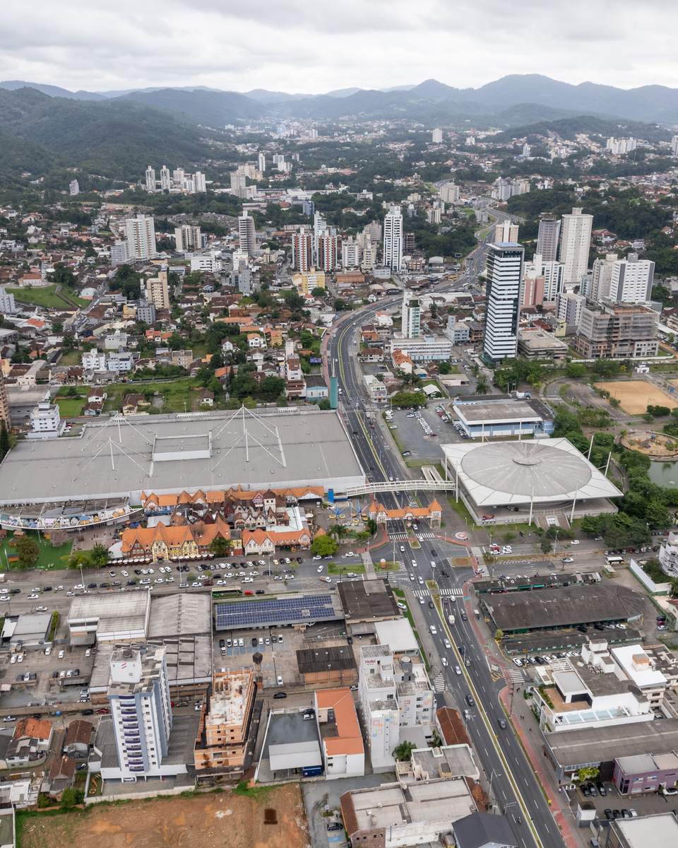 Vista aérea do Centro de Blumenau num dia nublado, com prédios, avenidas, os pavilhões do Parque Vila Germânica no meio e os morros verdes ao fundo.
