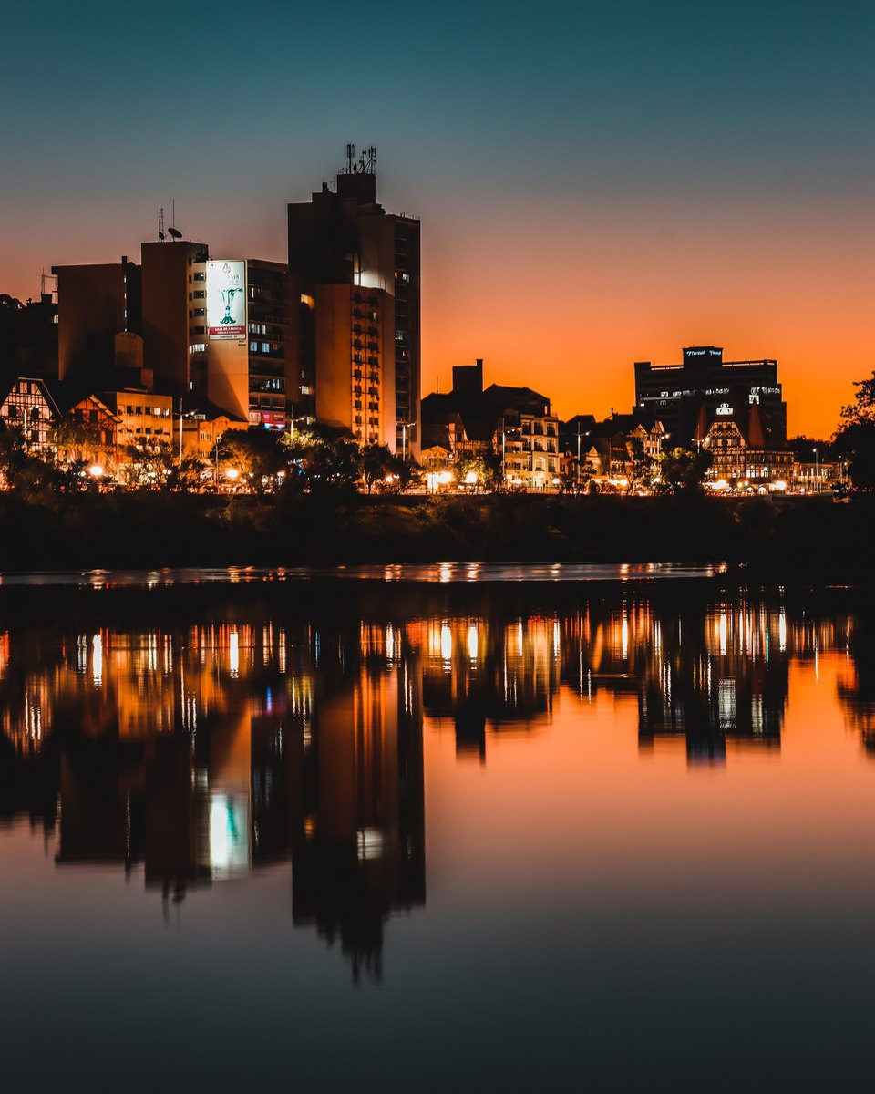 Prédios do Centro de Blumenau iluminados à noite e refletidos no rio Itajaí-Açu, com o céu alaranjado do entardecer.