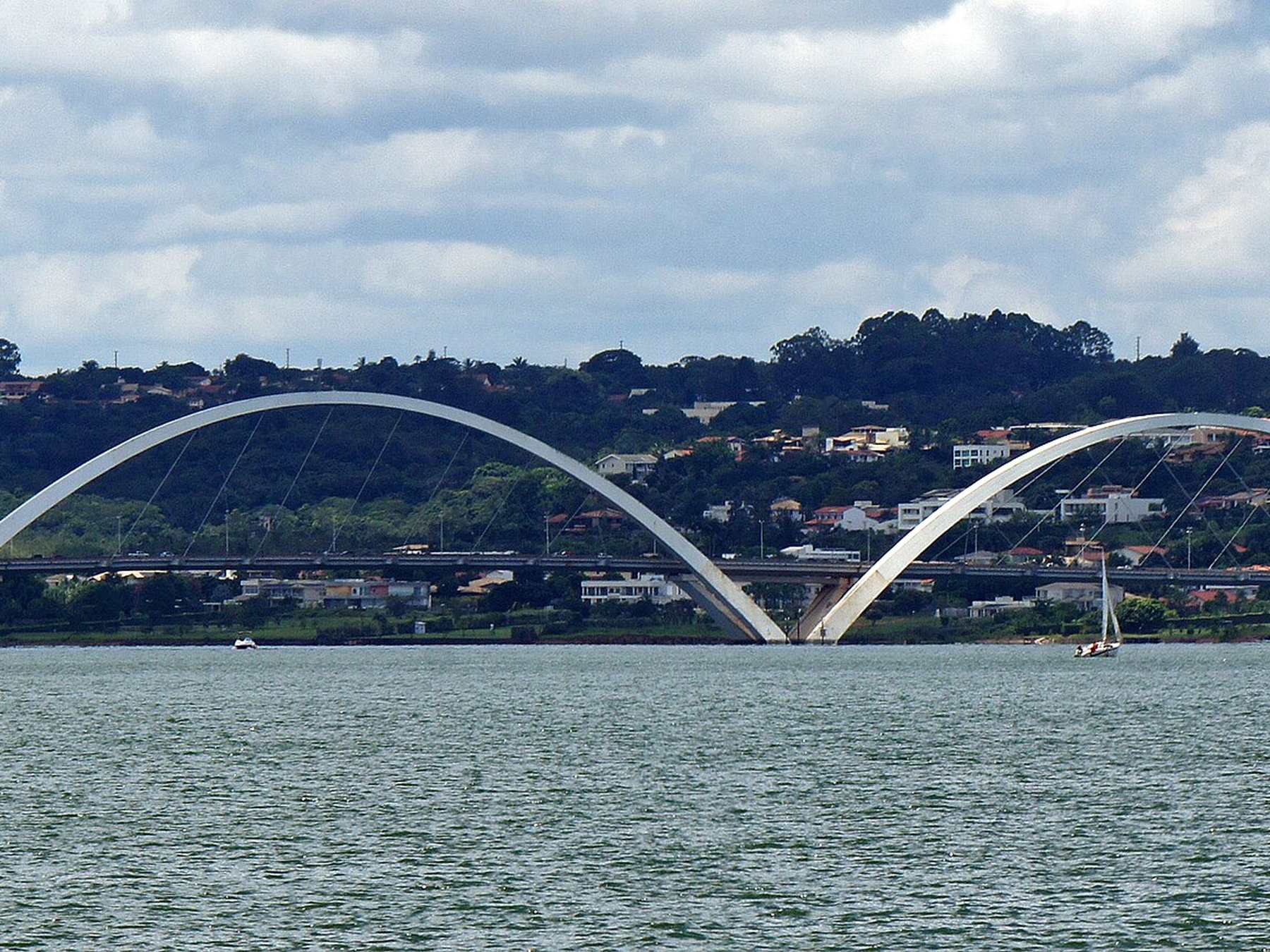 Panorâmica da Ponte Juscelino Kubitschek, em Brasília, com os três arcos de aço cruzando o Lago Paranoá e a cidade ao fundo, vista da margem oposta.