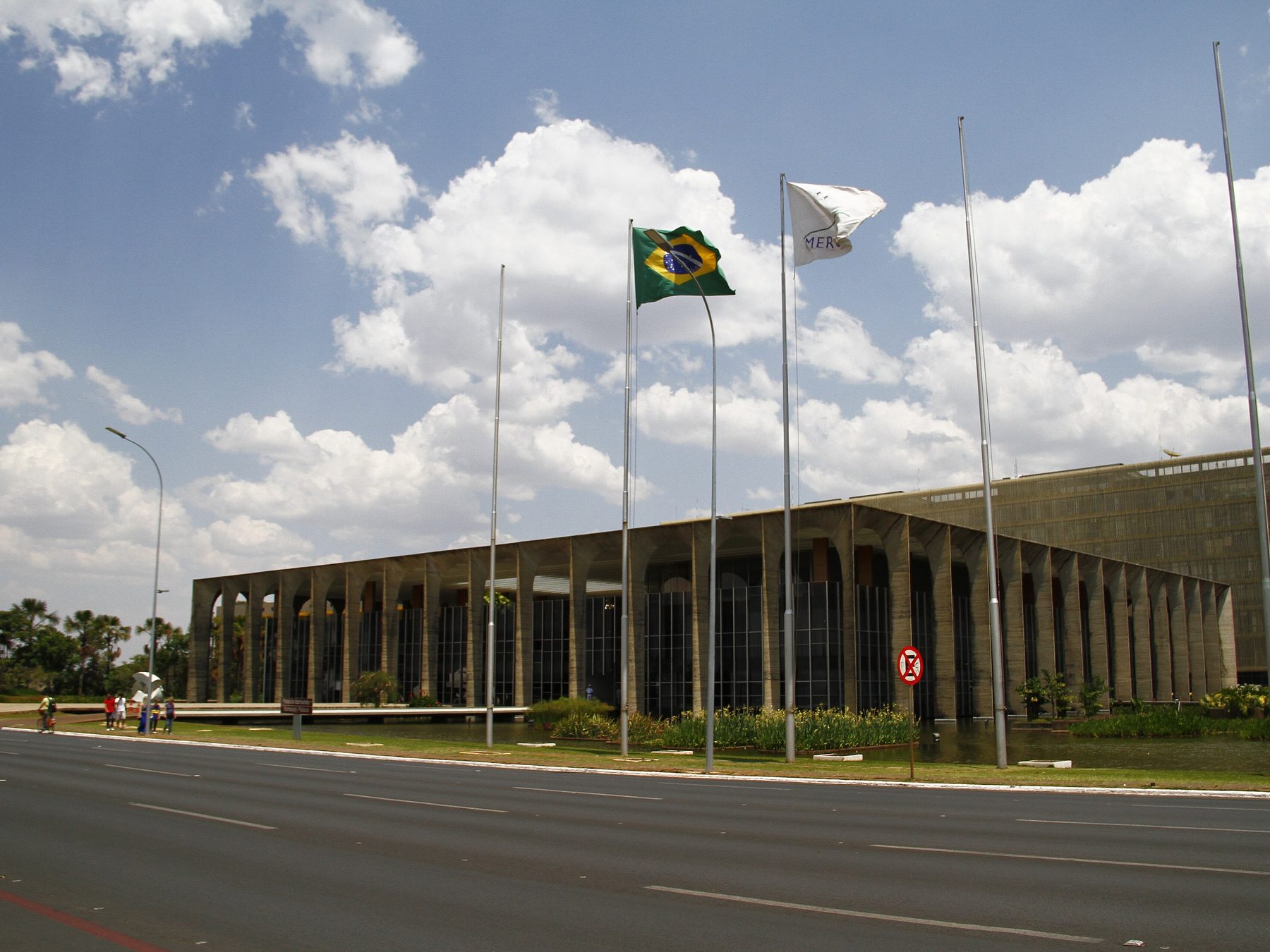 Palácio do Itamaraty, em Brasília, visto da avenida, com a fileira de arcos de concreto, o espelho d'água na base e mastros de bandeira em frente, sob céu de nuvens brancas.