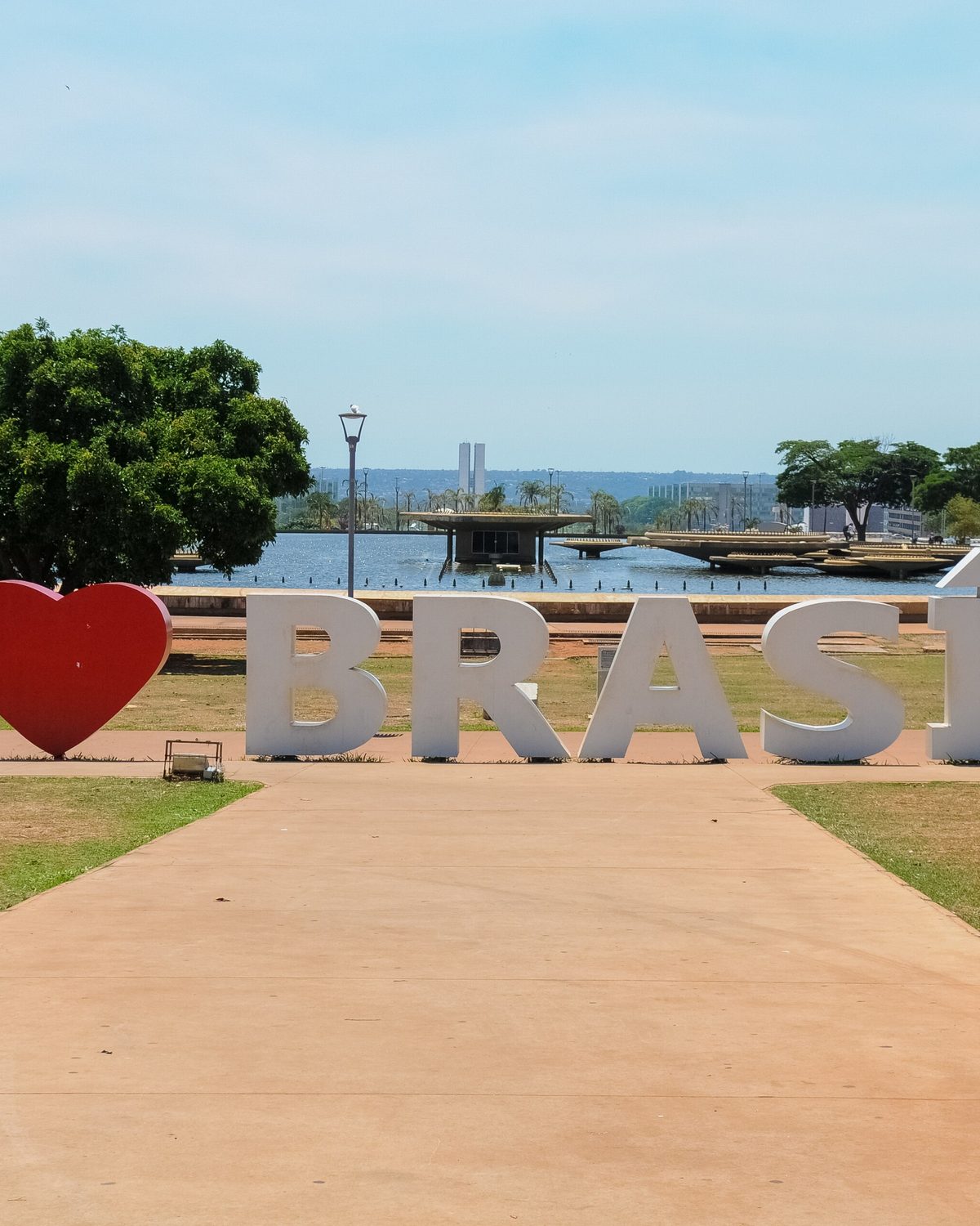 Letreiro com as palavras Eu amo Brasília em letras brancas e um coração vermelho, no gramado do Eixo Monumental, com a Fonte da Torre de TV e a cidade ao fundo, num dia de sol.