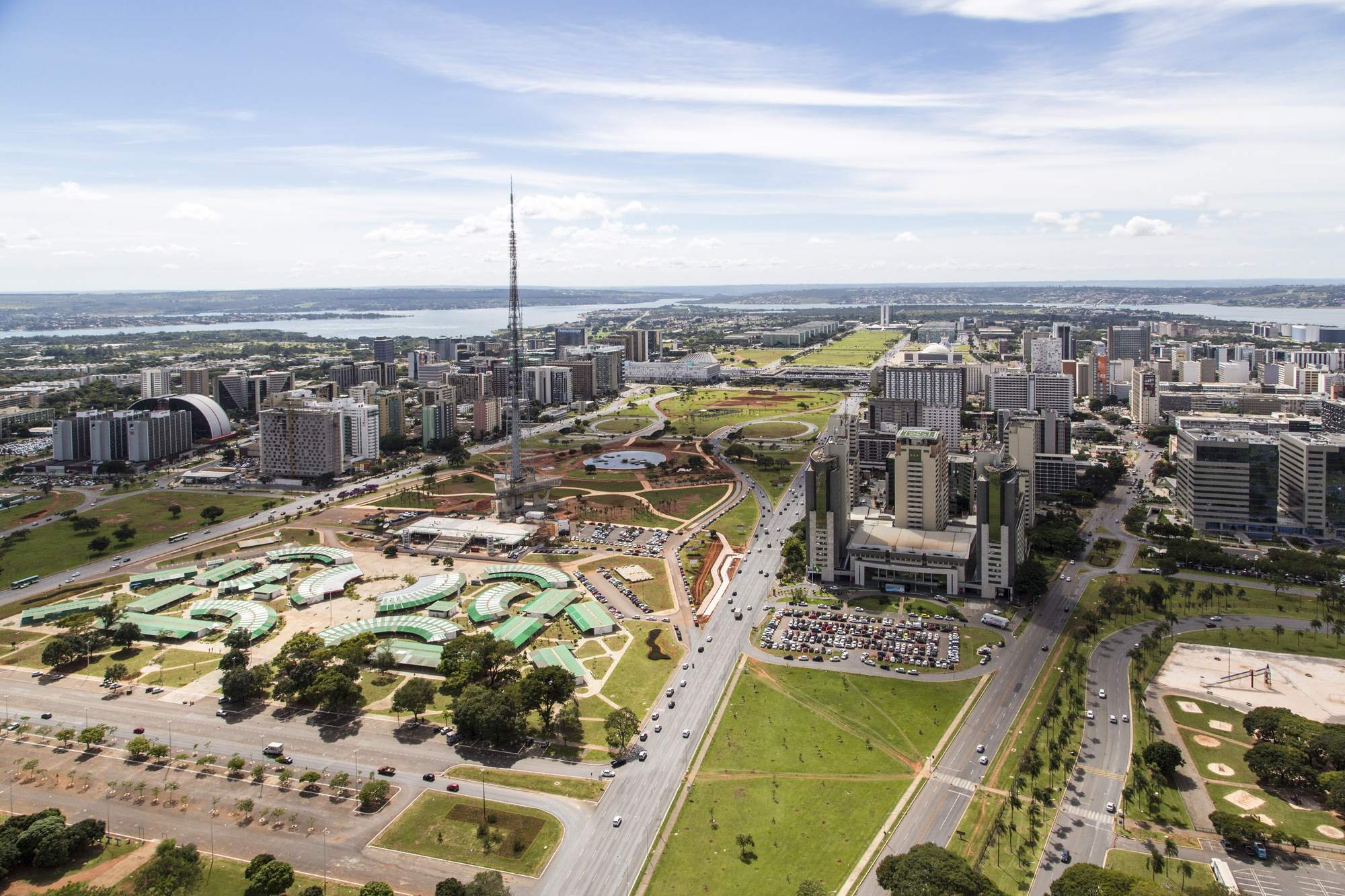 Vista aérea do Eixo Monumental de Brasília num dia de sol, com a Torre de TV no meio do gramado, as barracas da feira embaixo dela, as pistas em linha reta dos dois lados e o Lago Paranoá no horizonte.