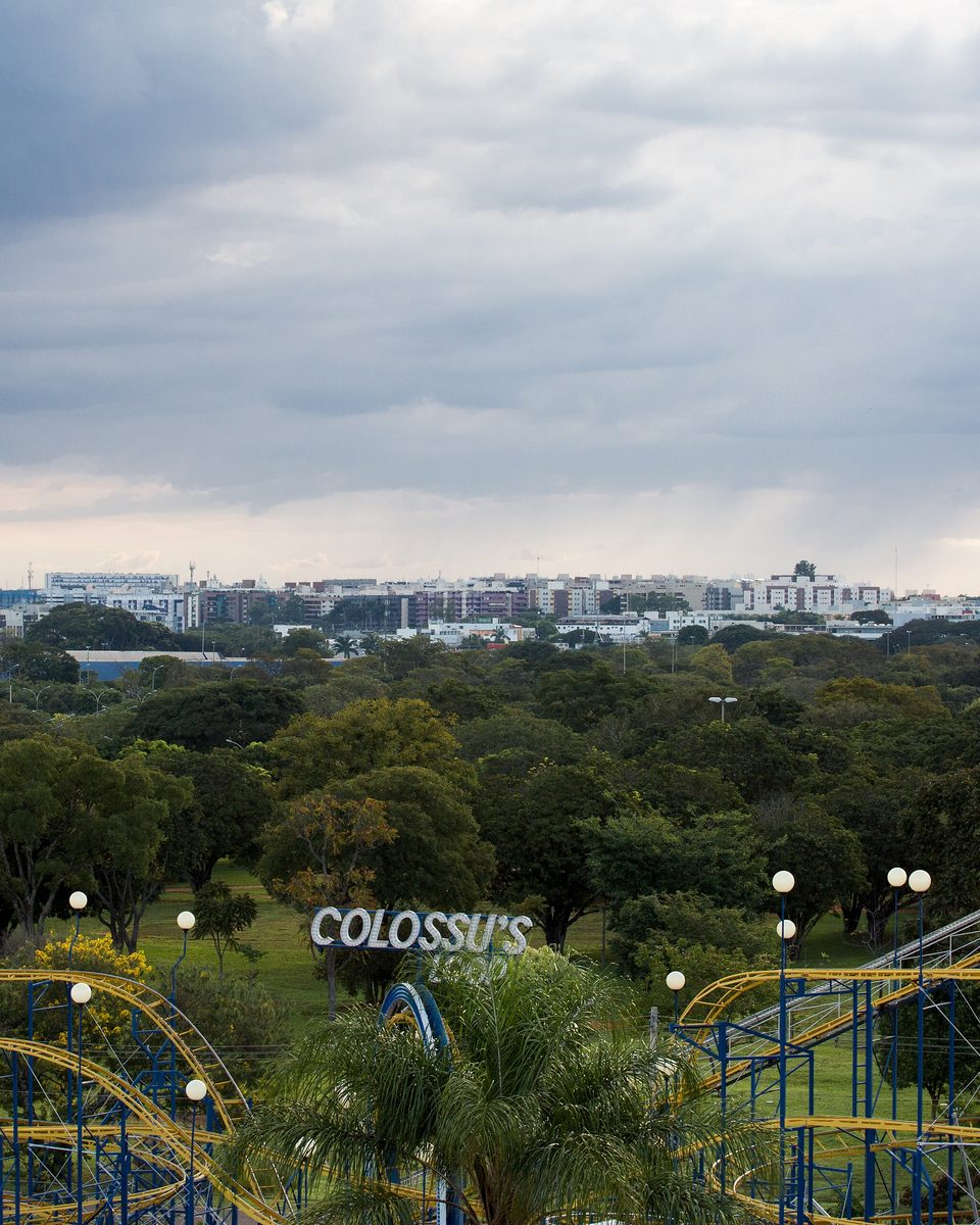 Vista do alto do Parque da Cidade Dona Sarah Kubitschek, na Asa Sul de Brasília, com a montanha-russa do parque de diversões em primeiro plano, a mata do parque ao redor e os prédios do Plano Piloto no horizonte.