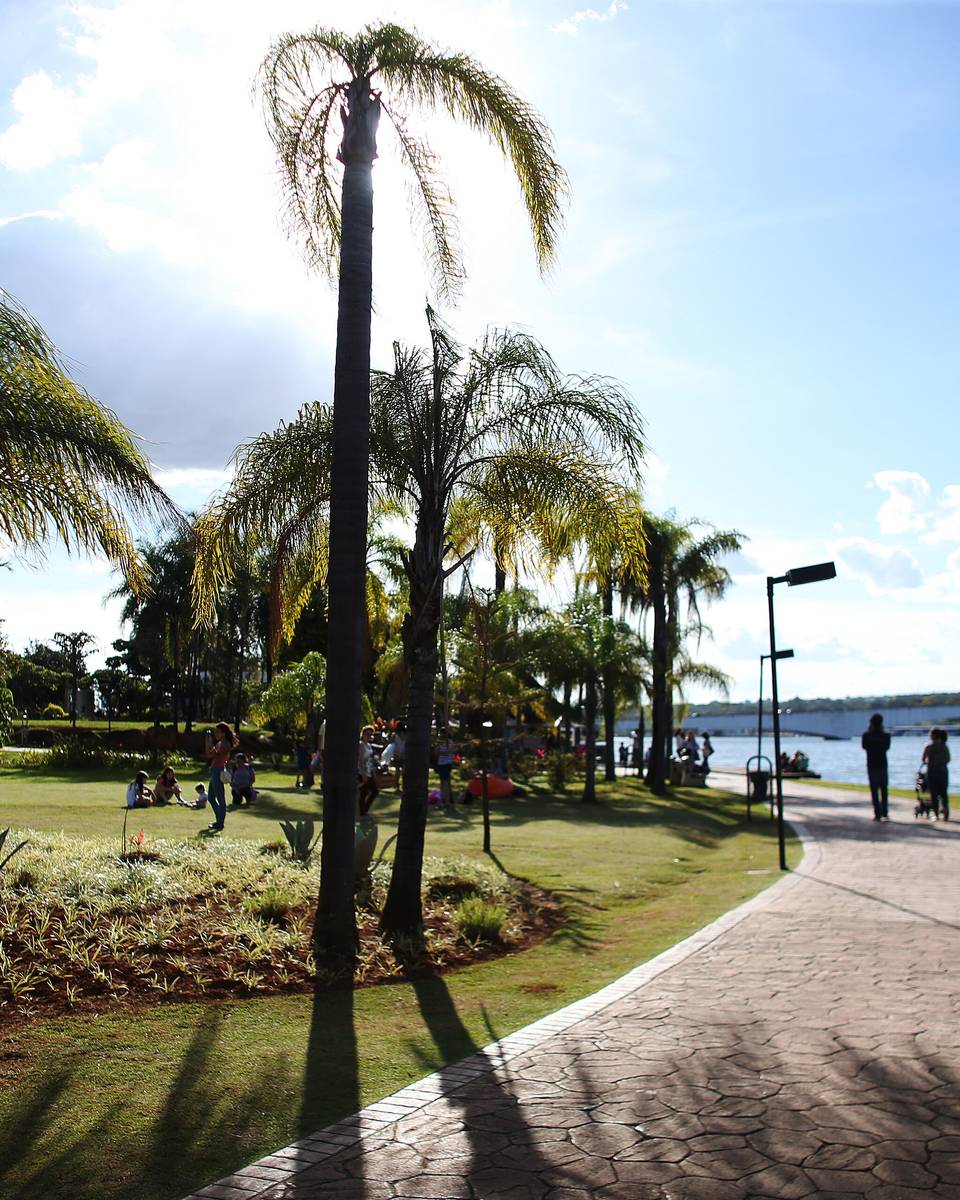 Calçadão do Pontão do Lago Sul, em Brasília, com palmeiras altas, gramado e o Lago Paranoá se abrindo ao lado, num fim de tarde de céu claro.